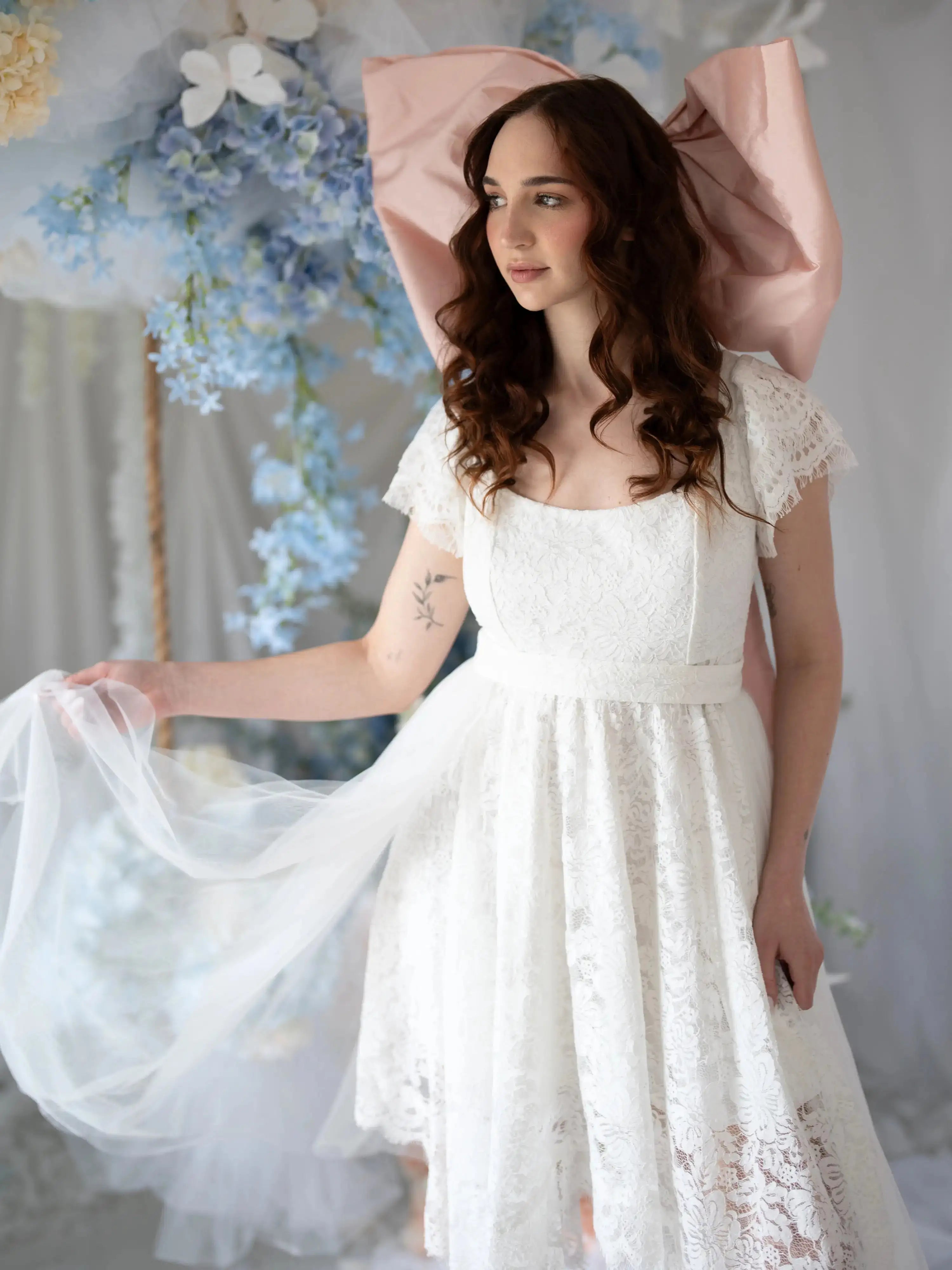 Romantic Belladonna Lace Dress with flutter sleeves and flowing train, styled in a floral wedding setting with pastel blue accents.