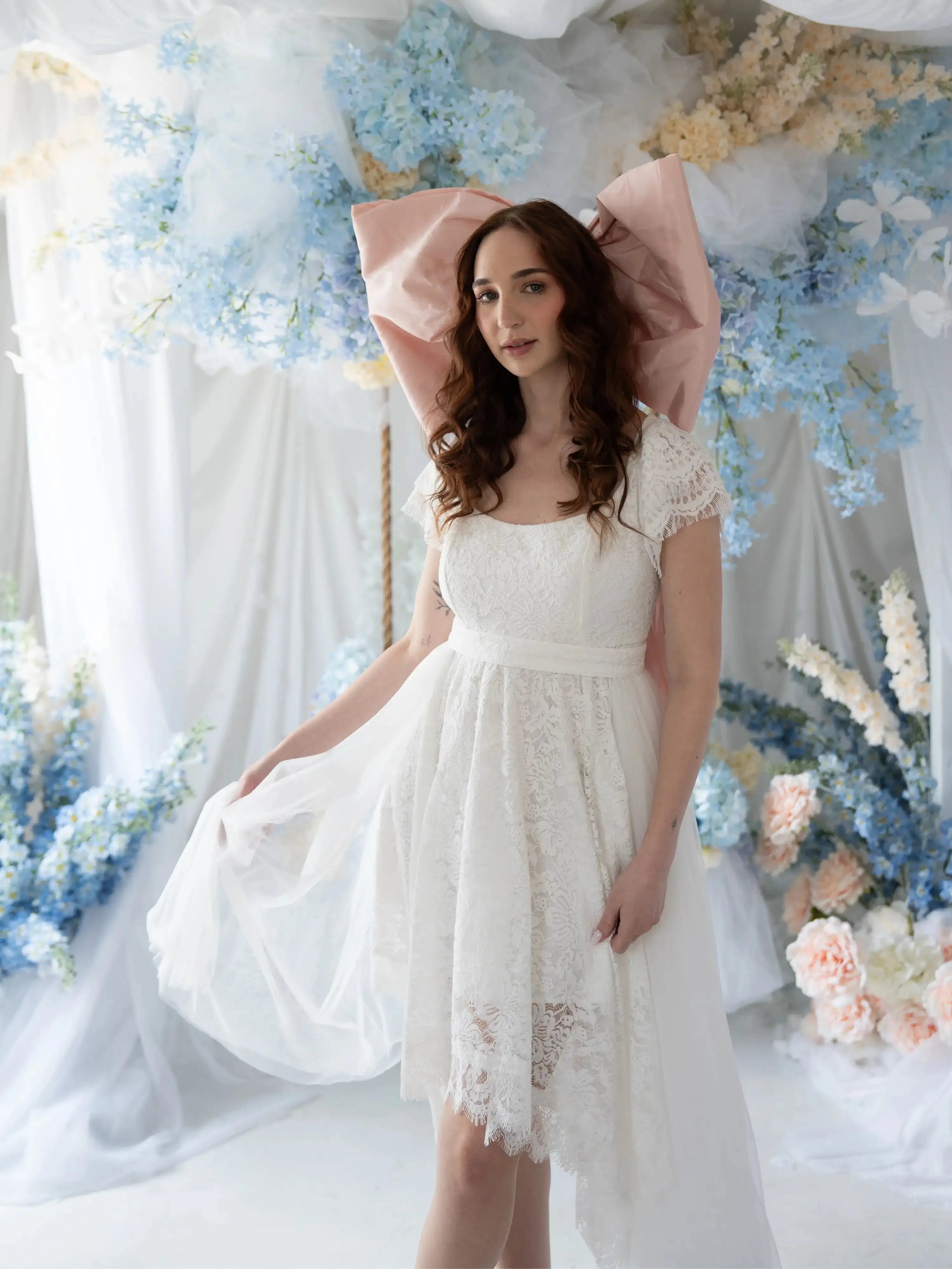 Belladonna Lace Dress styled in dreamy floral backdrop, showcasing lace bodice, soft sleeves, and flowing high-low hemline.