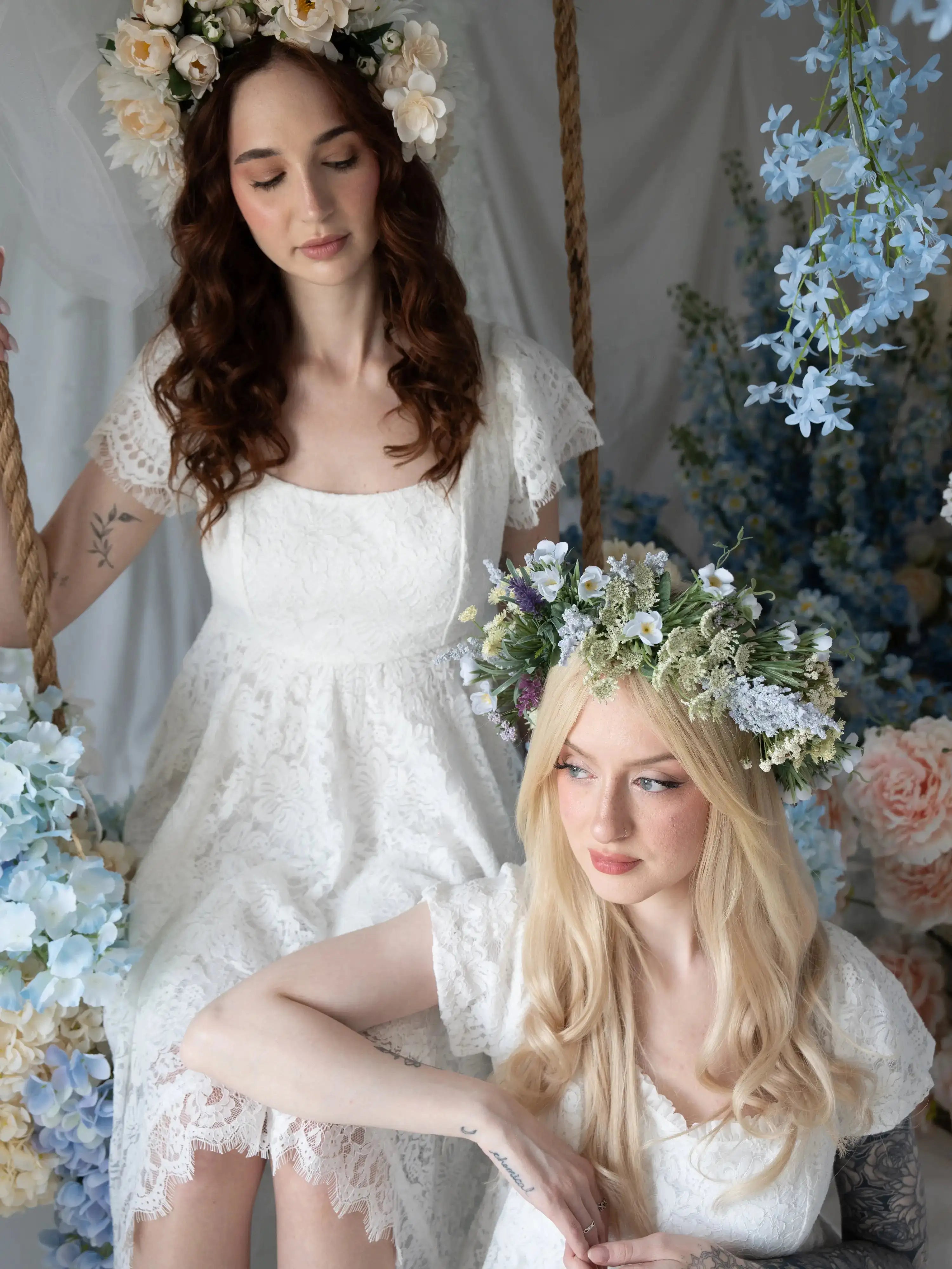 Two models in Belladonna Lace Dresses with floral crowns, seated on a swing surrounded by pastel wedding florals and lace details.