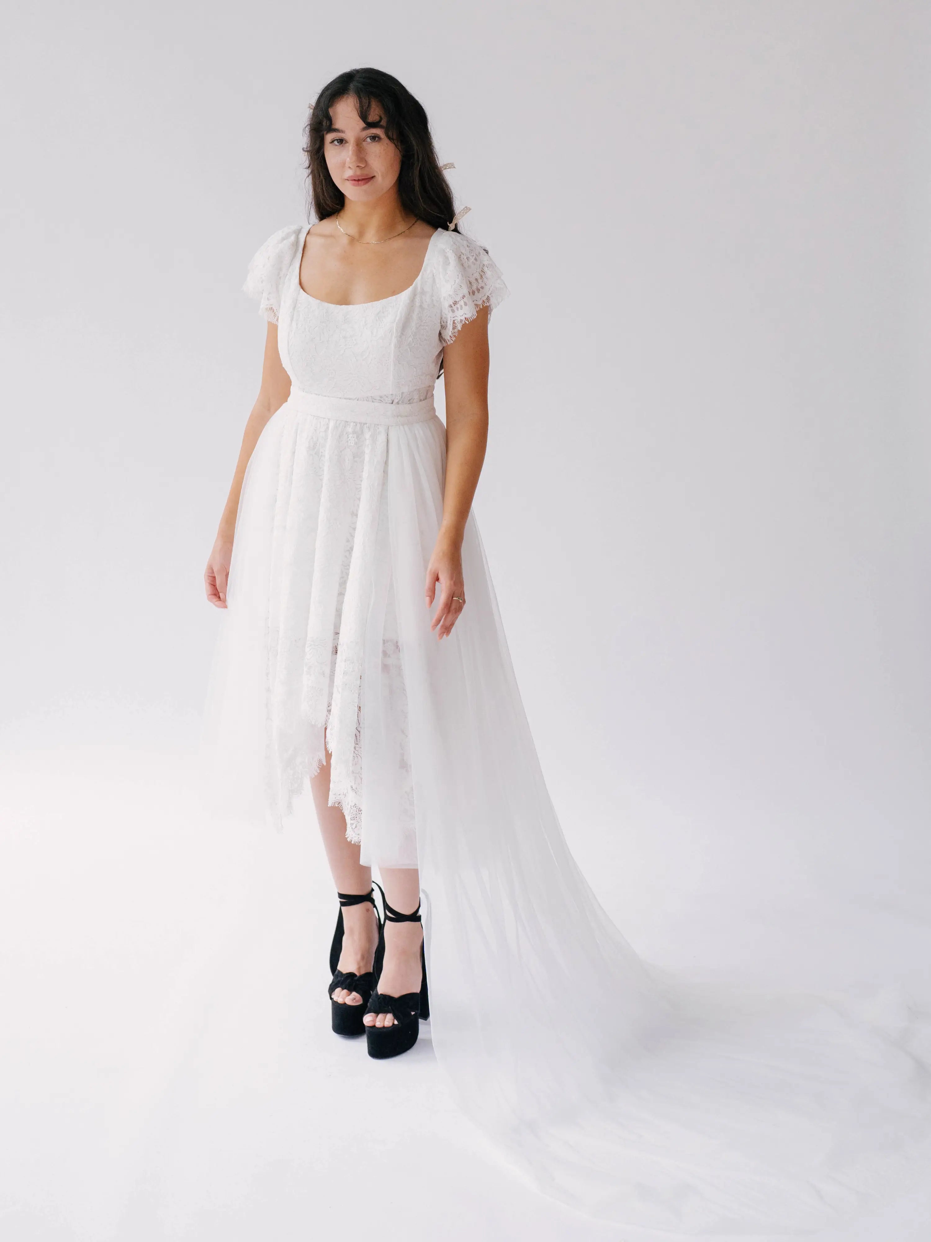 Belladonna Lace Dress midi bridal gown with high-low hem and trailing tulle train, modeled in studio with black platform heels.