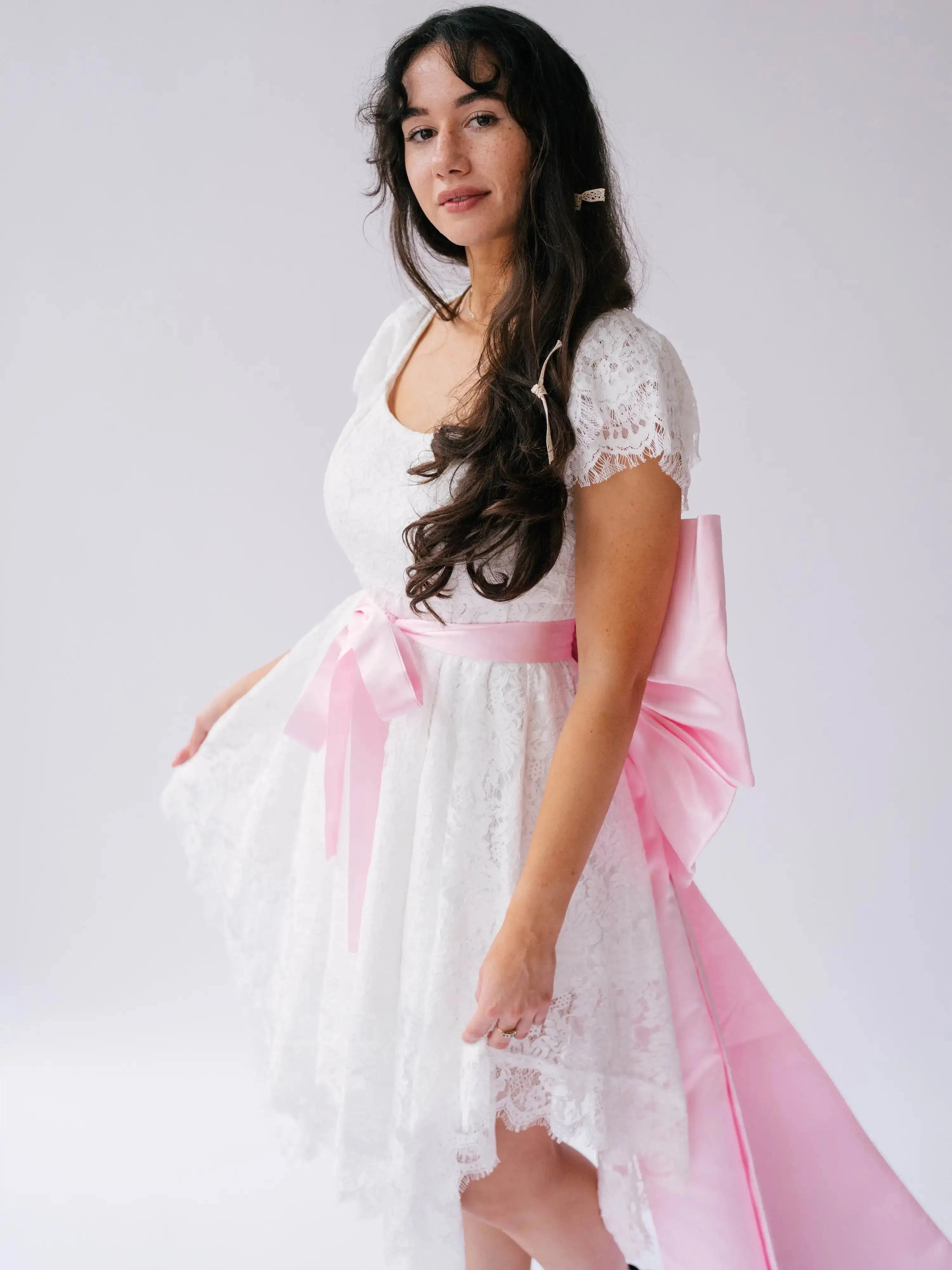Belladonna Lace Dress styled with pink satin bow sash, highlighting romantic bridal design with lace and high-low silhouette.
