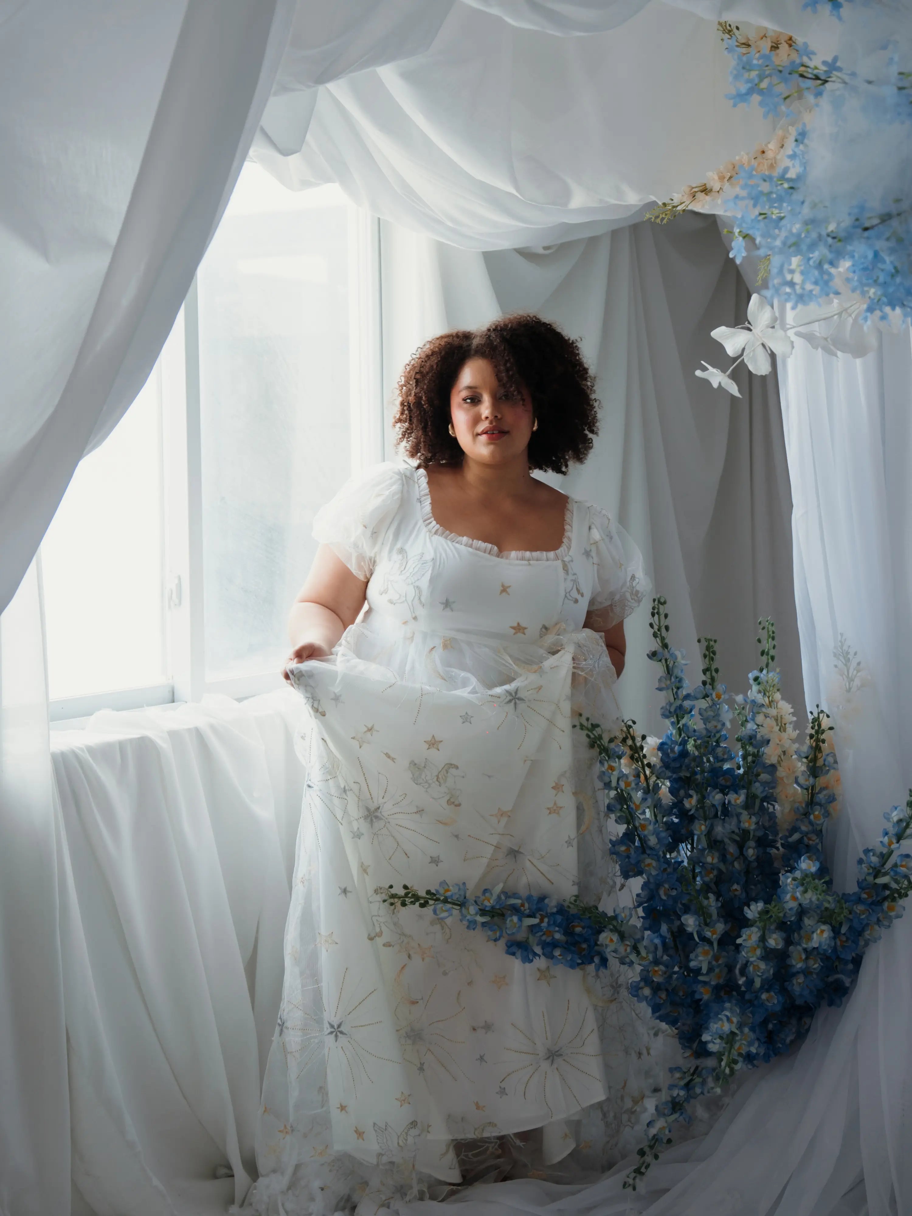 Woman in a white dress holding a floral arrangement in a decorated room with white curtains and blue flowers.