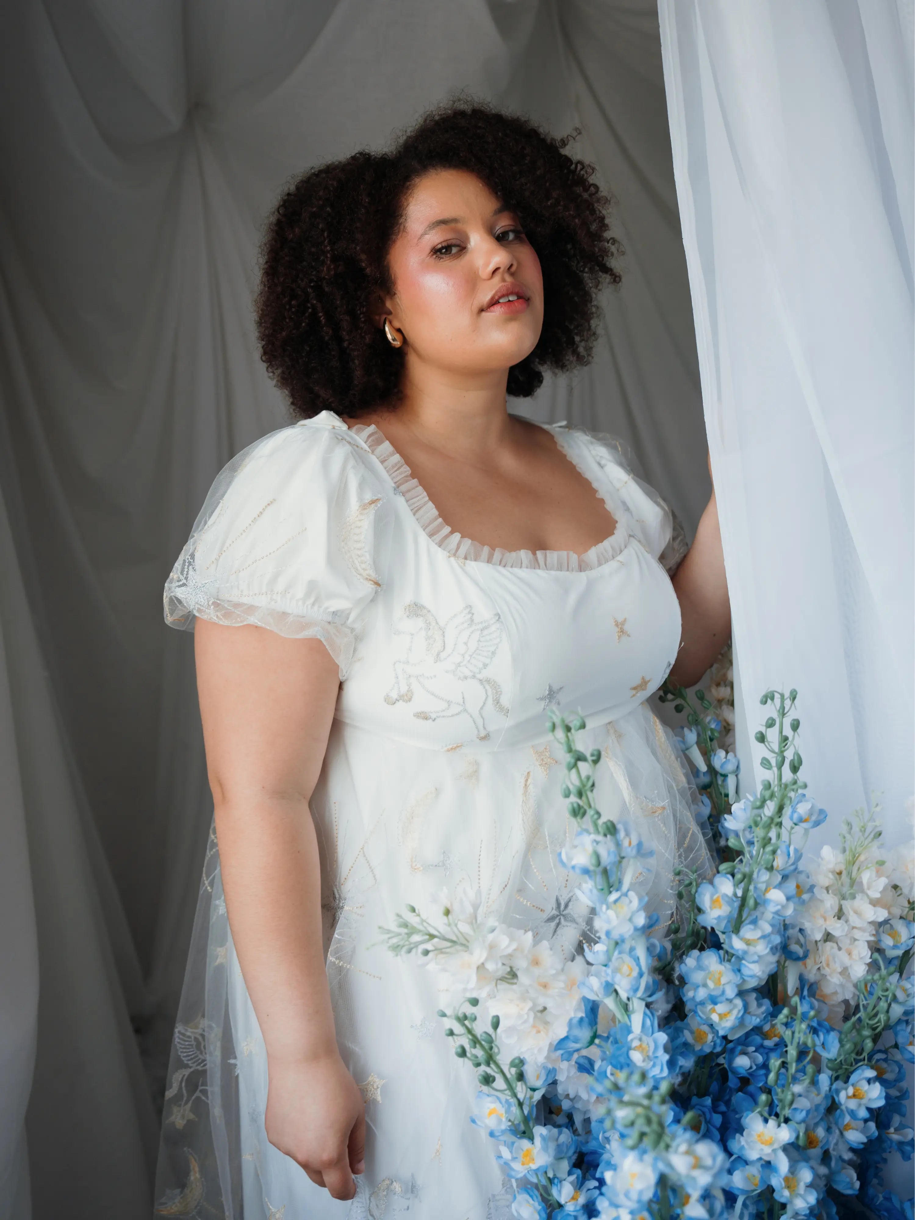 Woman in a white dress standing near a bouquet of flowers against a white curtain.