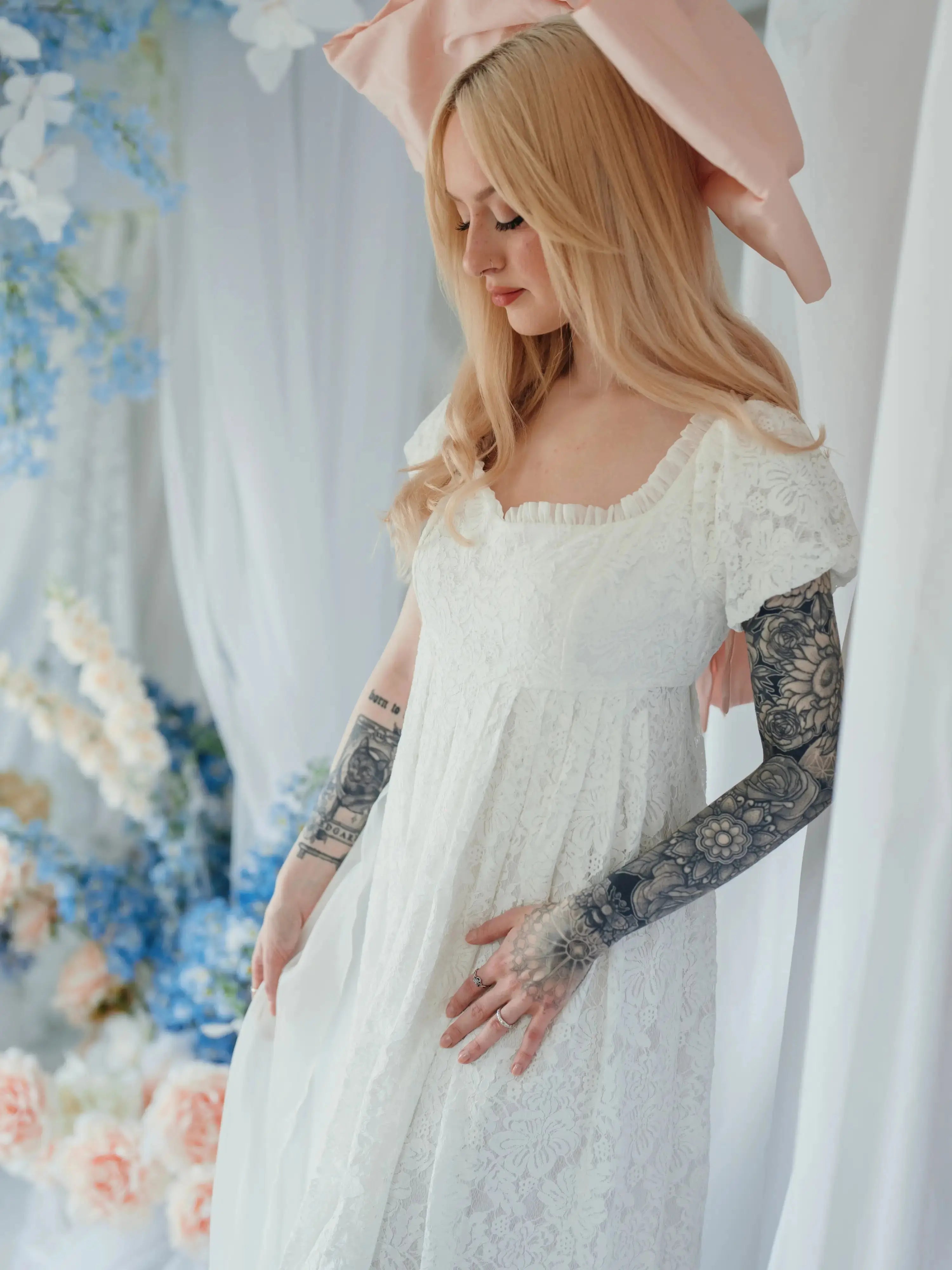 Dauphine Lace Gown with ruffled neckline and puff sleeves, styled with pink bow in a floral wedding backdrop.