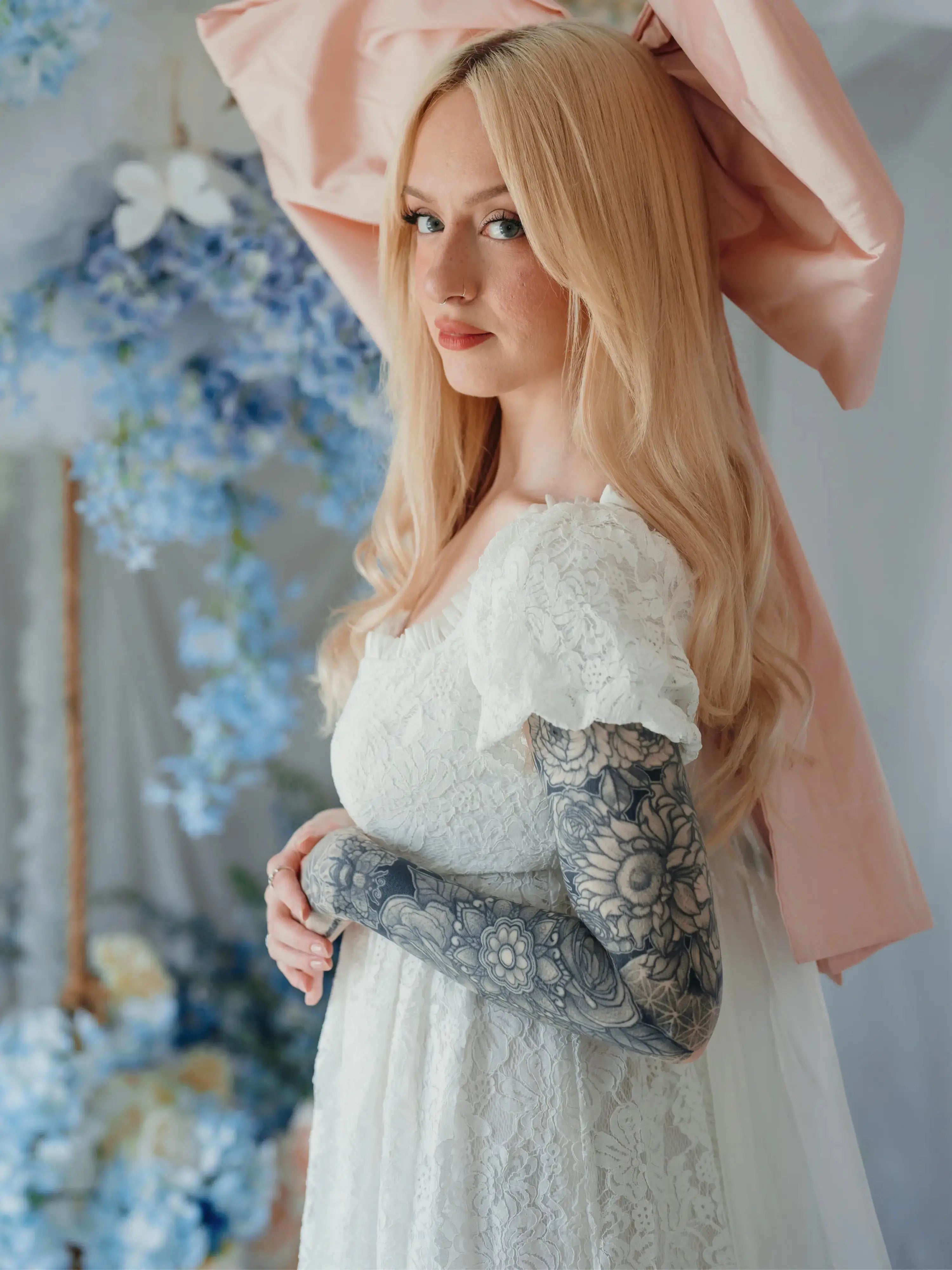 Side profile of model in Dauphine Lace Gown with puff sleeves and pink bow, surrounded by pastel floral wedding decor.