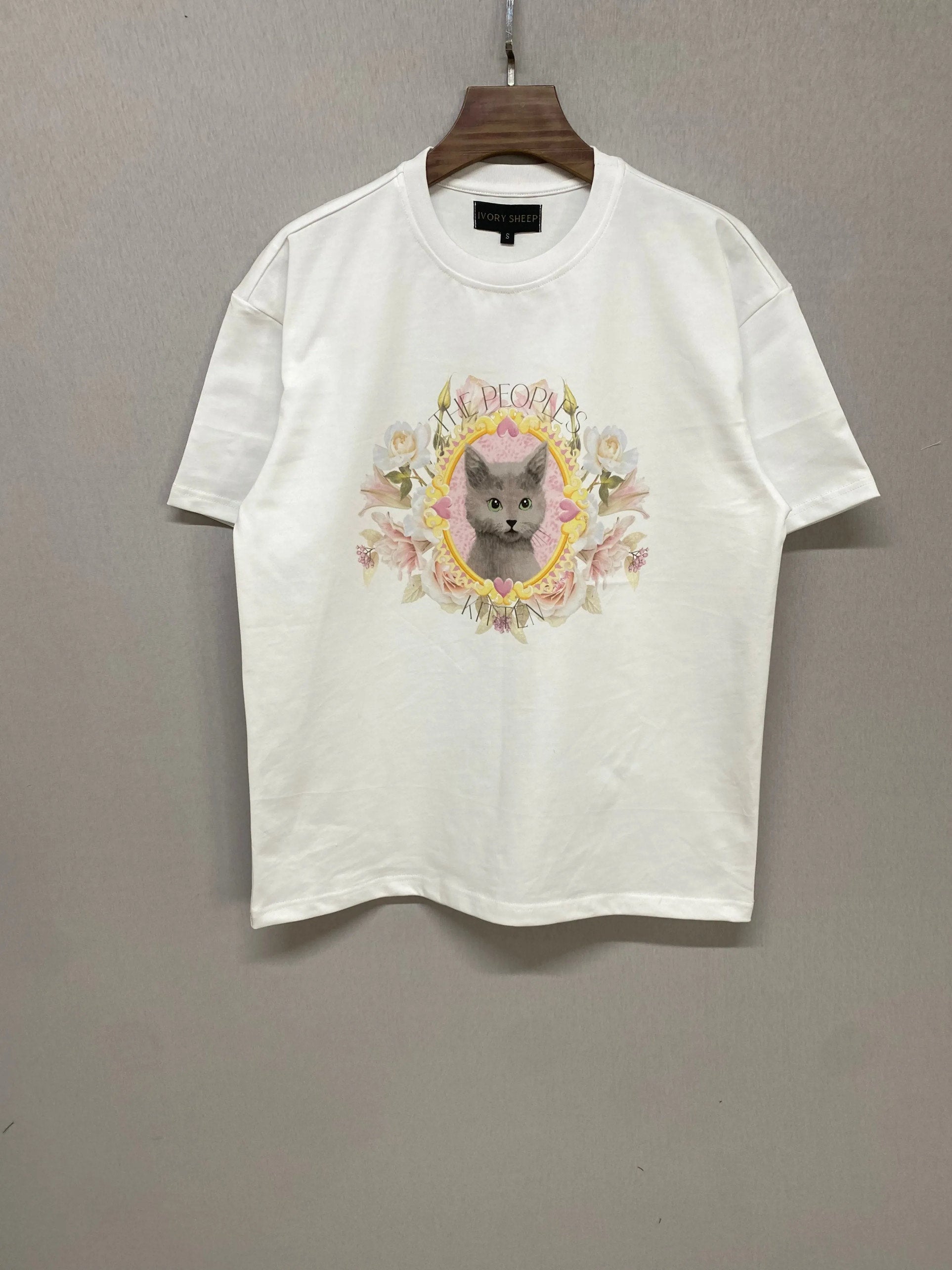 Flat lay of The Princess Kitten Tee on hanger, showing the full front design with pastel kitten artwork and floral details on white cotton.