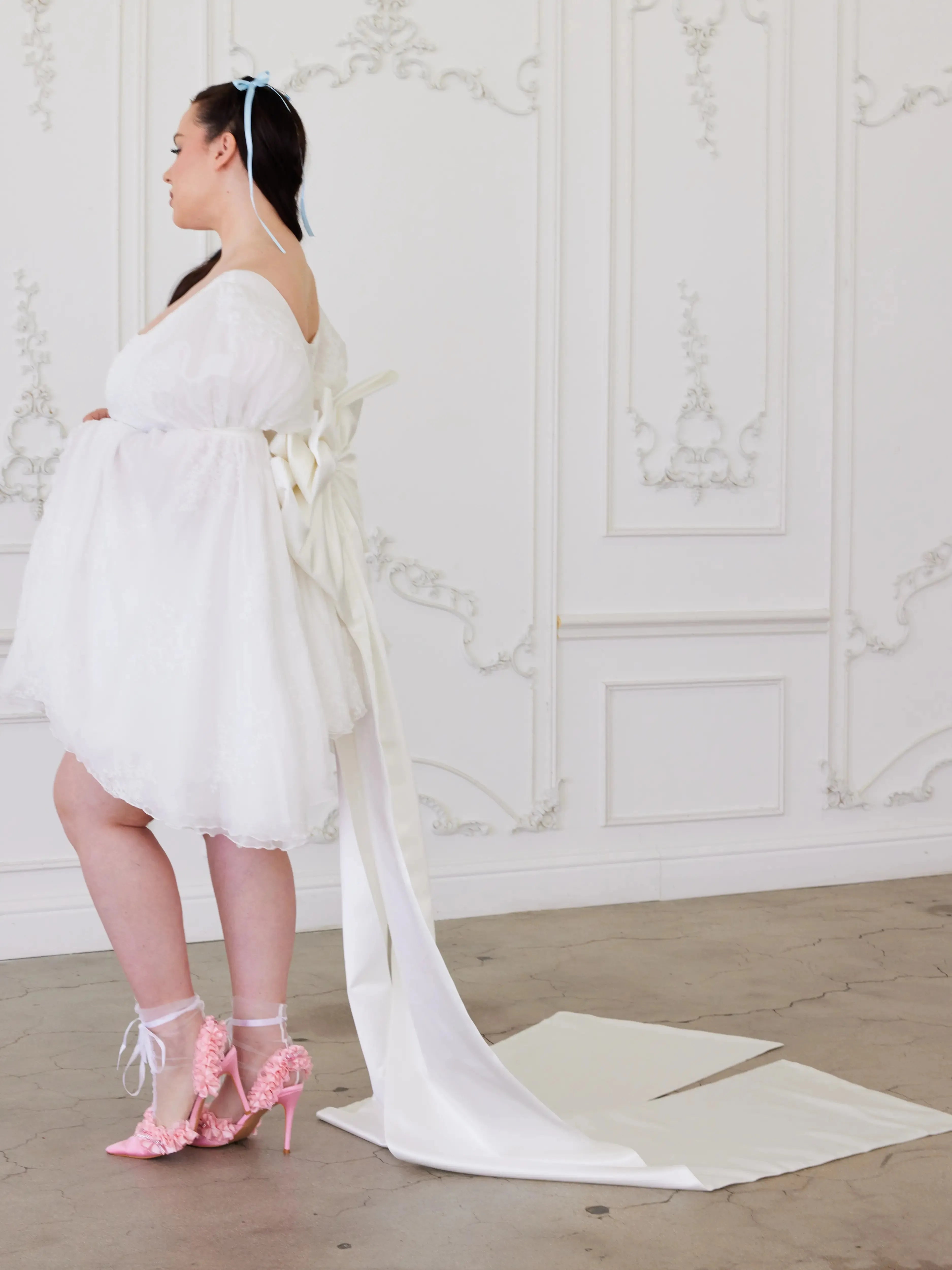 Full-length view of the Ivory Bridal Grand Bow Belt with extended satin train, styled with a short white bridal dress and pink heels.