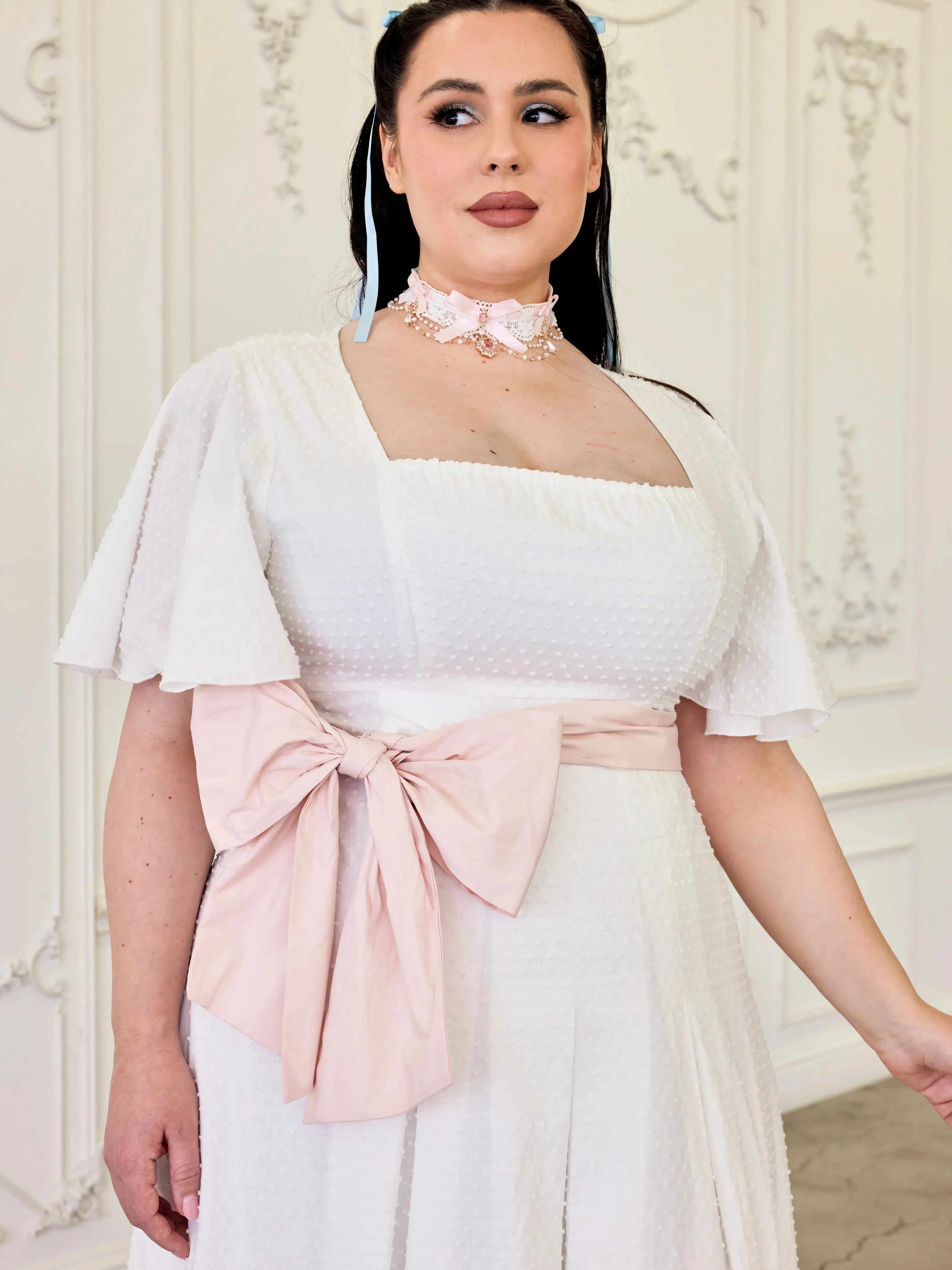 The Ivory Cloud Cotton Day Dress with soft pink waist bow and flutter sleeves, showcasing textured Swiss dot fabric for a vintage-inspired look.