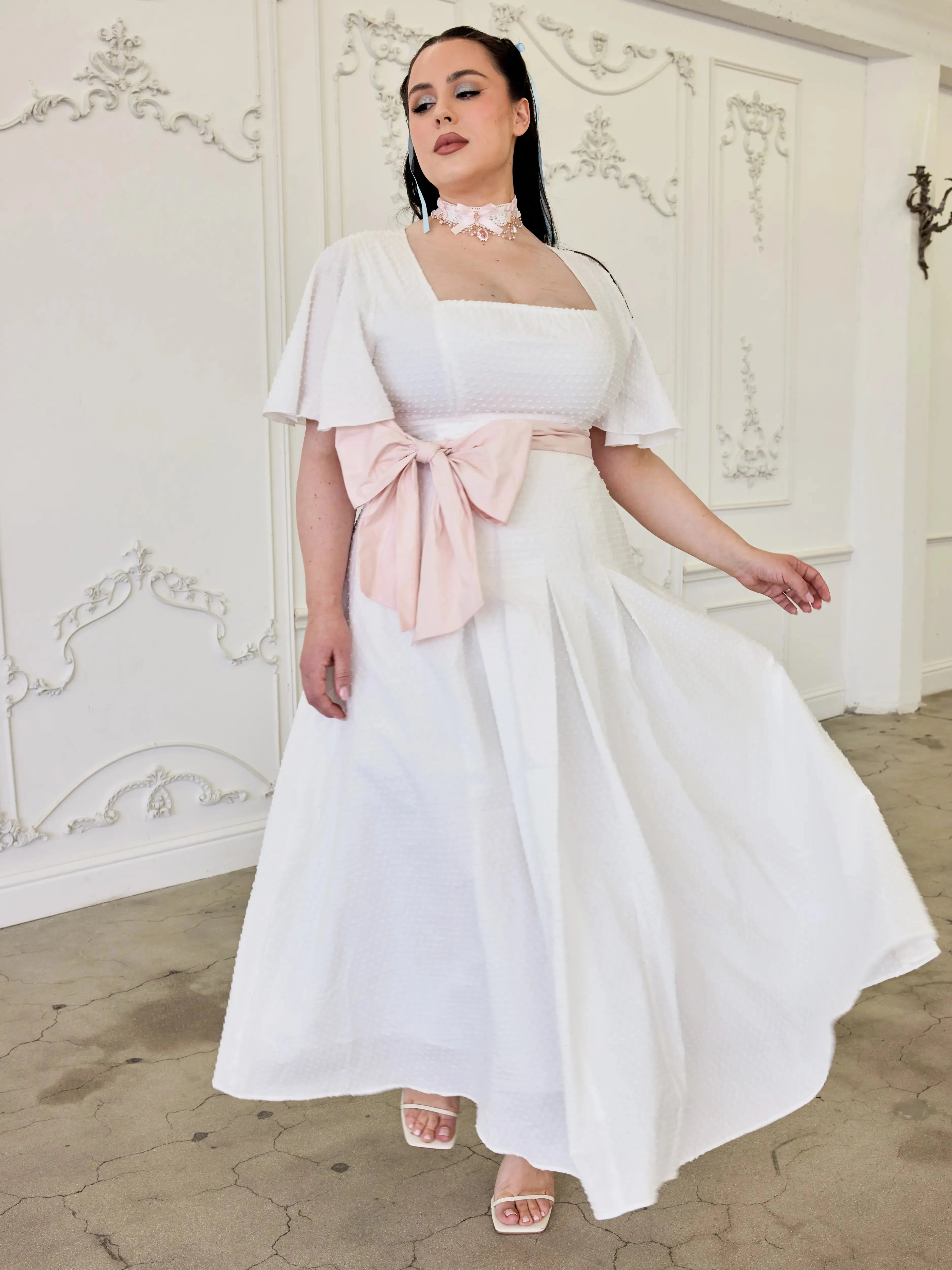 The Ivory Cloud Cotton Day Dress with pink sash and flutter sleeves, styled for summer weddings or romantic outings. Swiss dot texture adds elegance.