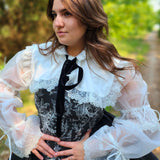Stylish Ciela Shrug Top featuring lace trim, puffed sleeves, and a black bow tie overtop The Rhiannon Corset Dress, worn by woman in the forest for a fairytale fashion look.