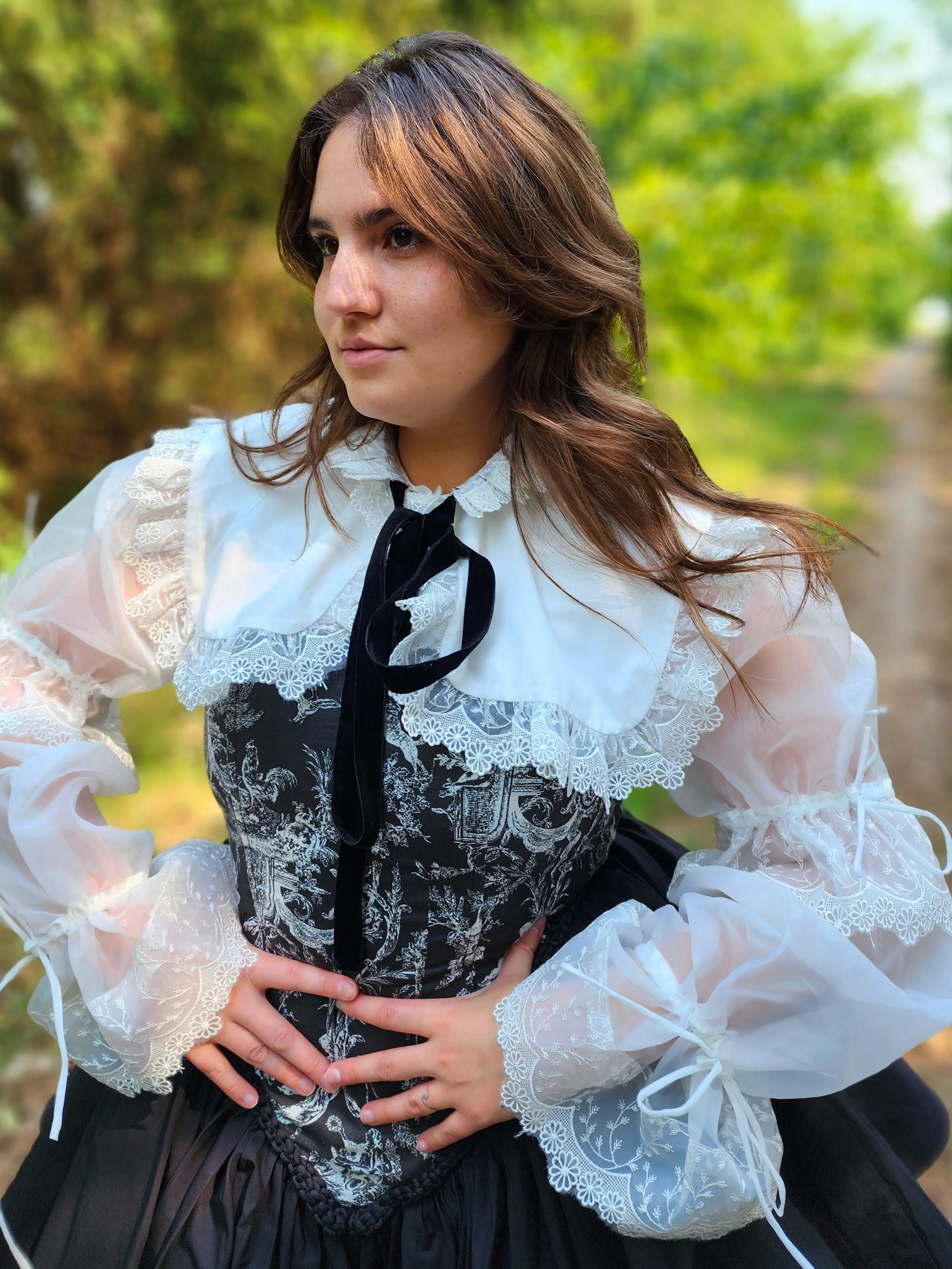 Stylish Ciela Shrug Top featuring lace trim, puffed sleeves, and a black bow tie overtop The Rhiannon Corset Dress, worn by woman in the forest for a fairytale fashion look.