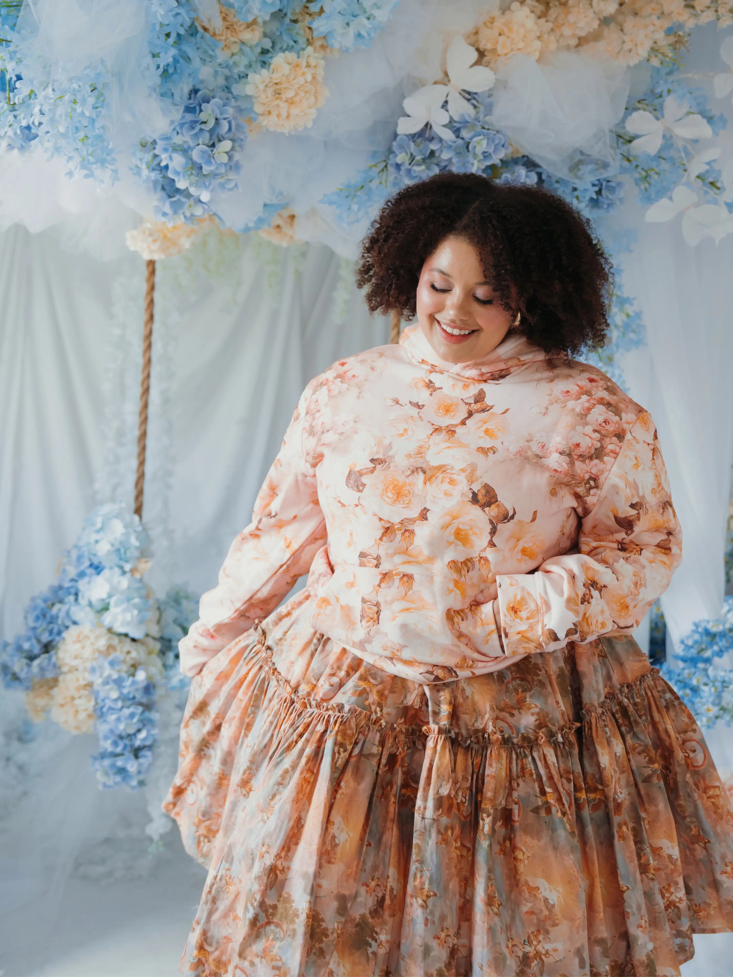 The Twilight Bloom Sleeper Hoodie paired with a floral skirt, modeled in a pastel floral backdrop. Oversized fit ideal for comfort and charm.
