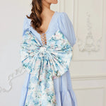 the Wildbloom Grand Bow Belt styled on a light blue dress, featuring an oversized floral satin bow with long trailing ribbons for romantic flair.