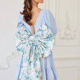the Wildbloom Grand Bow Belt styled on a light blue dress, featuring an oversized floral satin bow with long trailing ribbons for romantic flair.