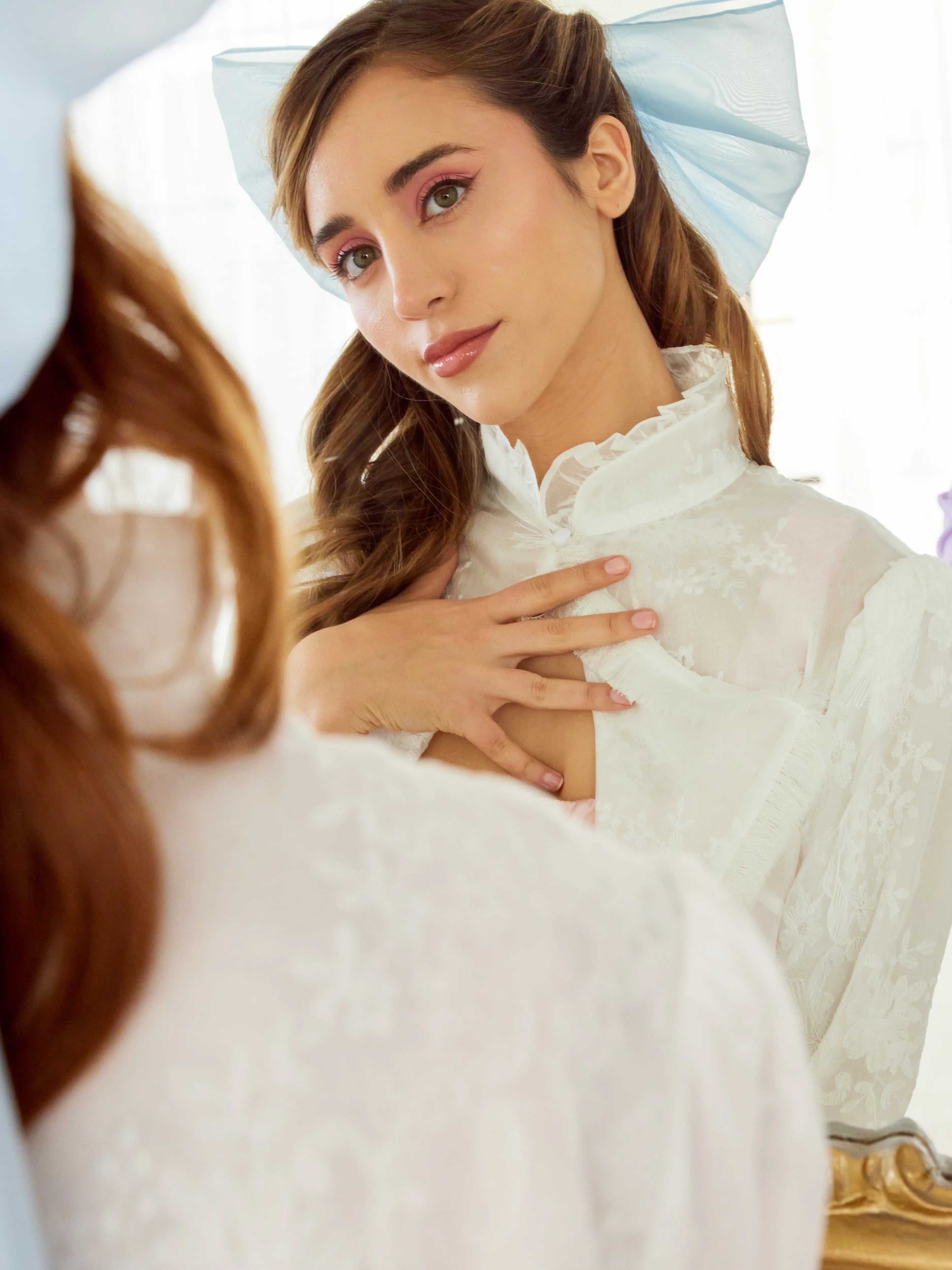 Caylee wearing the Alice Blue Grand Hair Bow styled with romantic waves and delicate lace attire, reflecting in a vintage mirror for a whimsical touch.