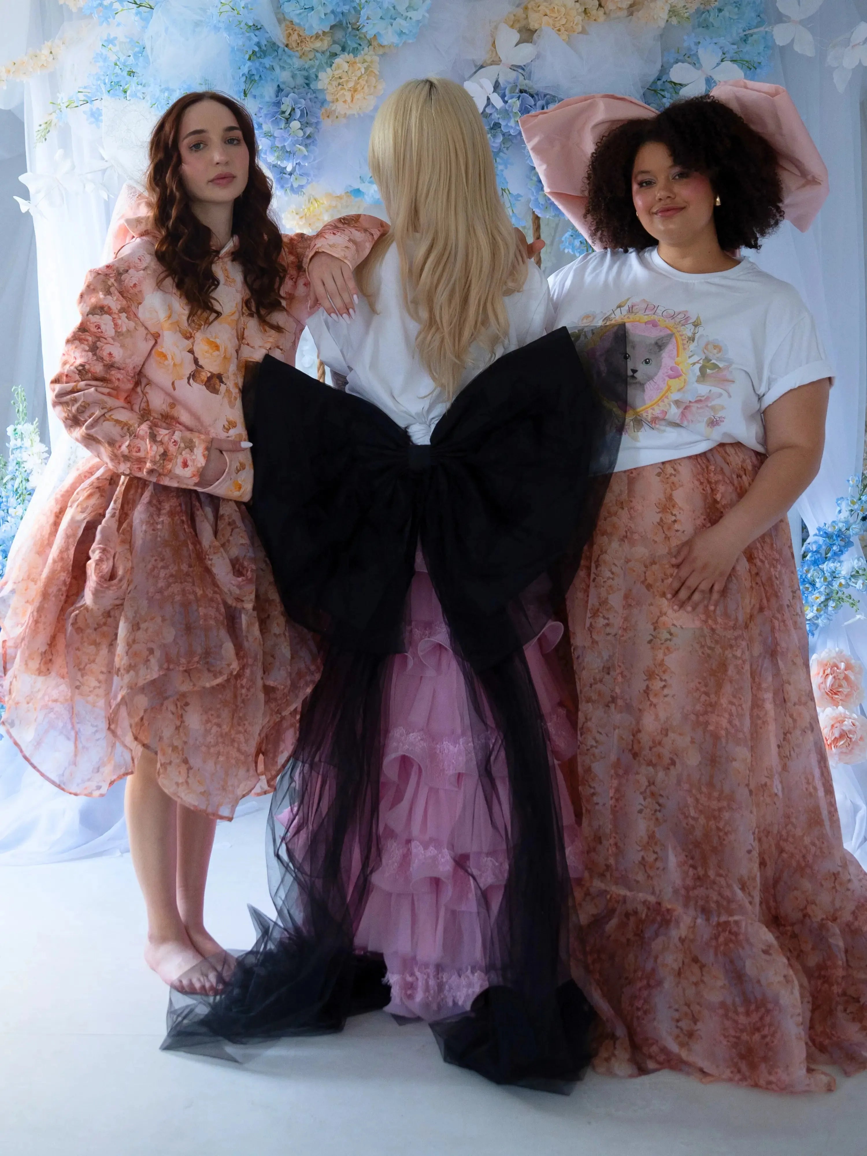The Black Swan Grand Bow Belt adds bold elegance to a whimsical group look, paired with floral dresses and pastel tones in a fairytale setting.