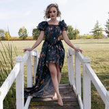 A woman in a dark autumn printed taffeta dress standing barefoot on a wooden bridge in a field