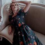 A woman wearing a still life printed taffeta dress sitting on an antique beige armchair with a window in the background