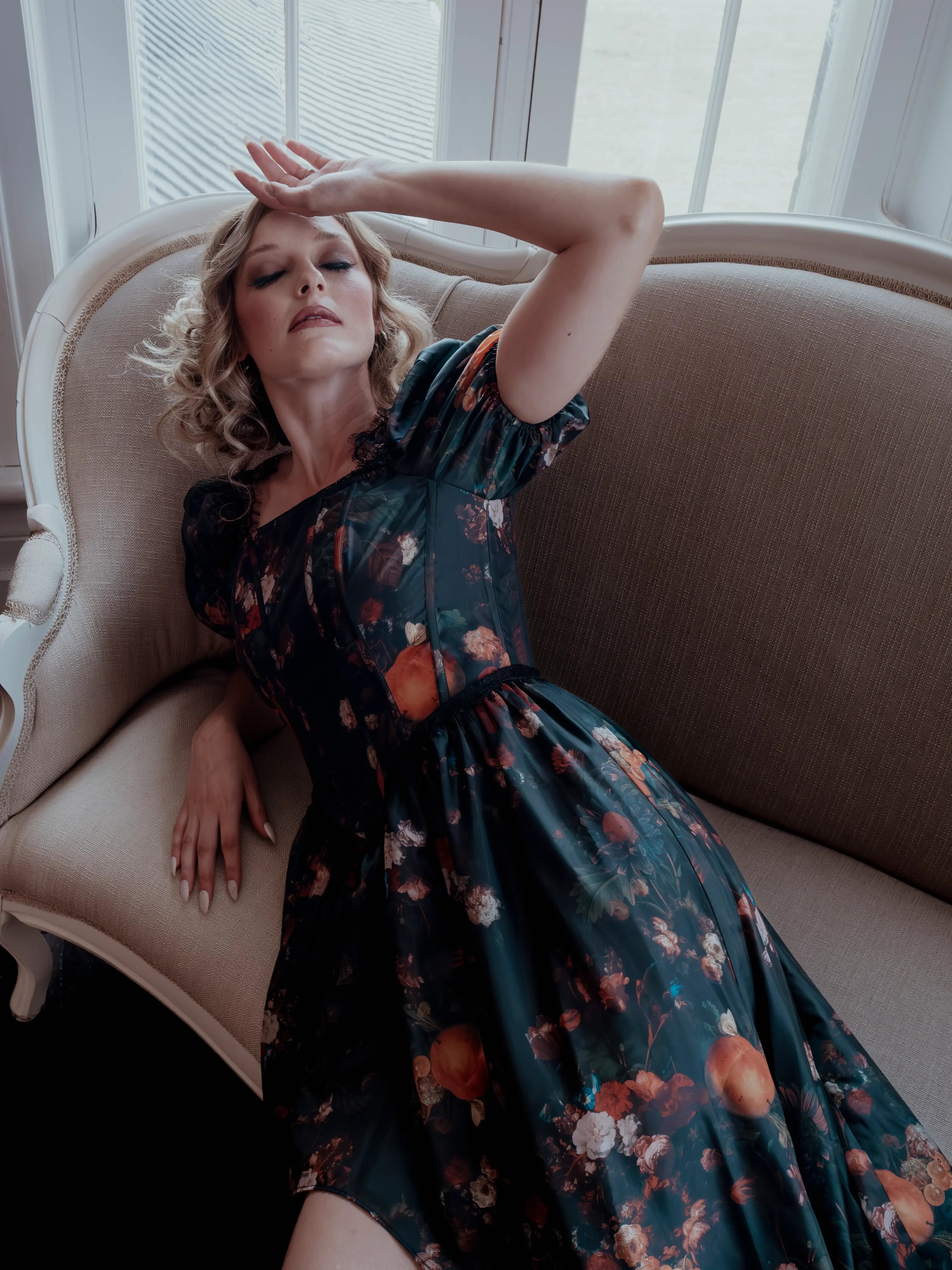 A woman wearing a still life printed taffeta dress sitting on an antique beige armchair with a window in the background