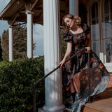 A woman wearing a still life printed taffeta dress standing on a porch with columns and greenery.
