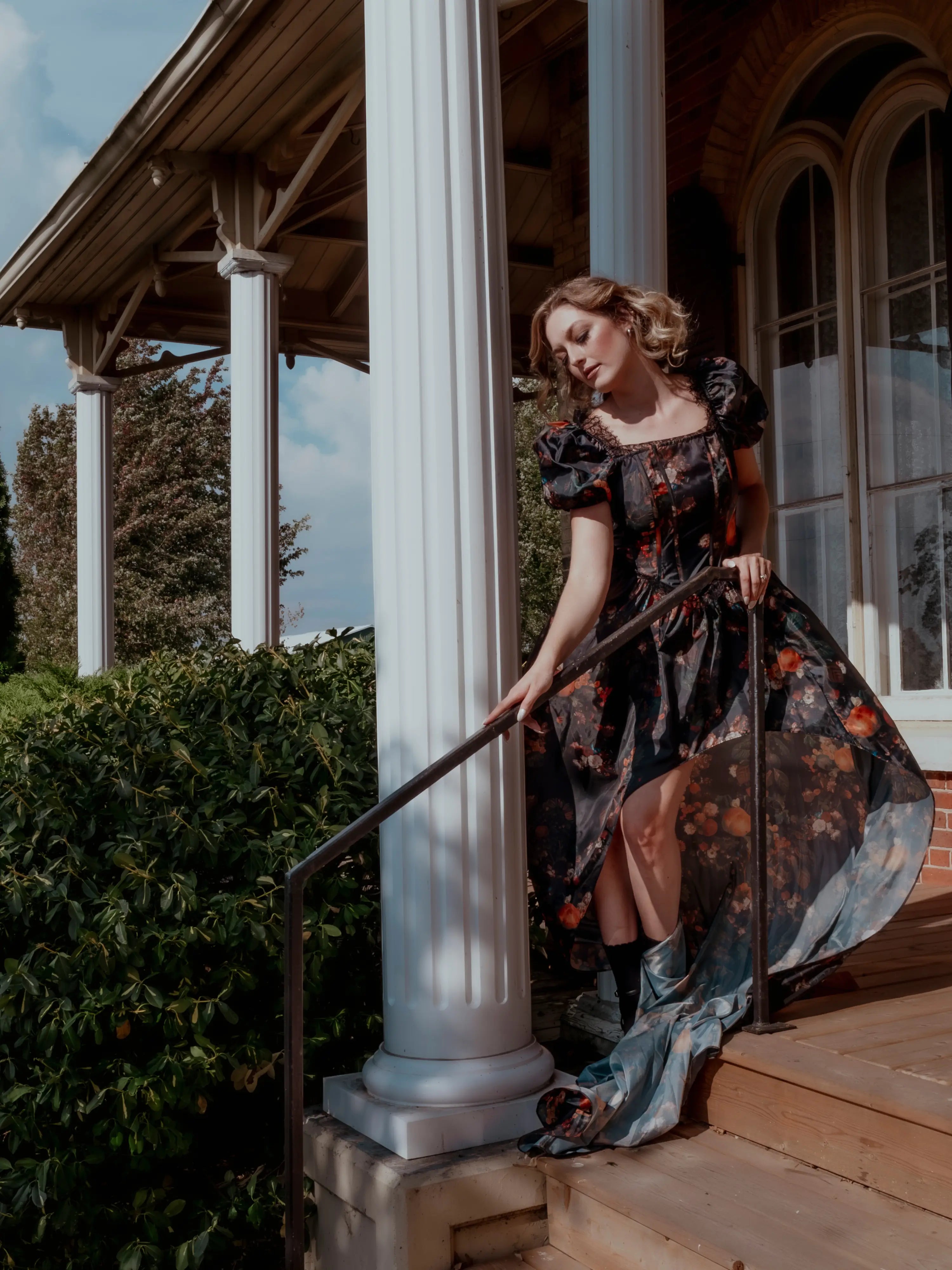 A woman wearing a still life printed taffeta dress standing on a porch with columns and greenery.