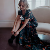 A woman wearing the still life Charlotte dress is sitting on the couch.