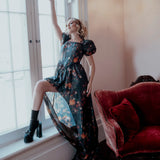 A woman wearing the still life printed taffeta dress is standing by a window with a red couch in the foreground.