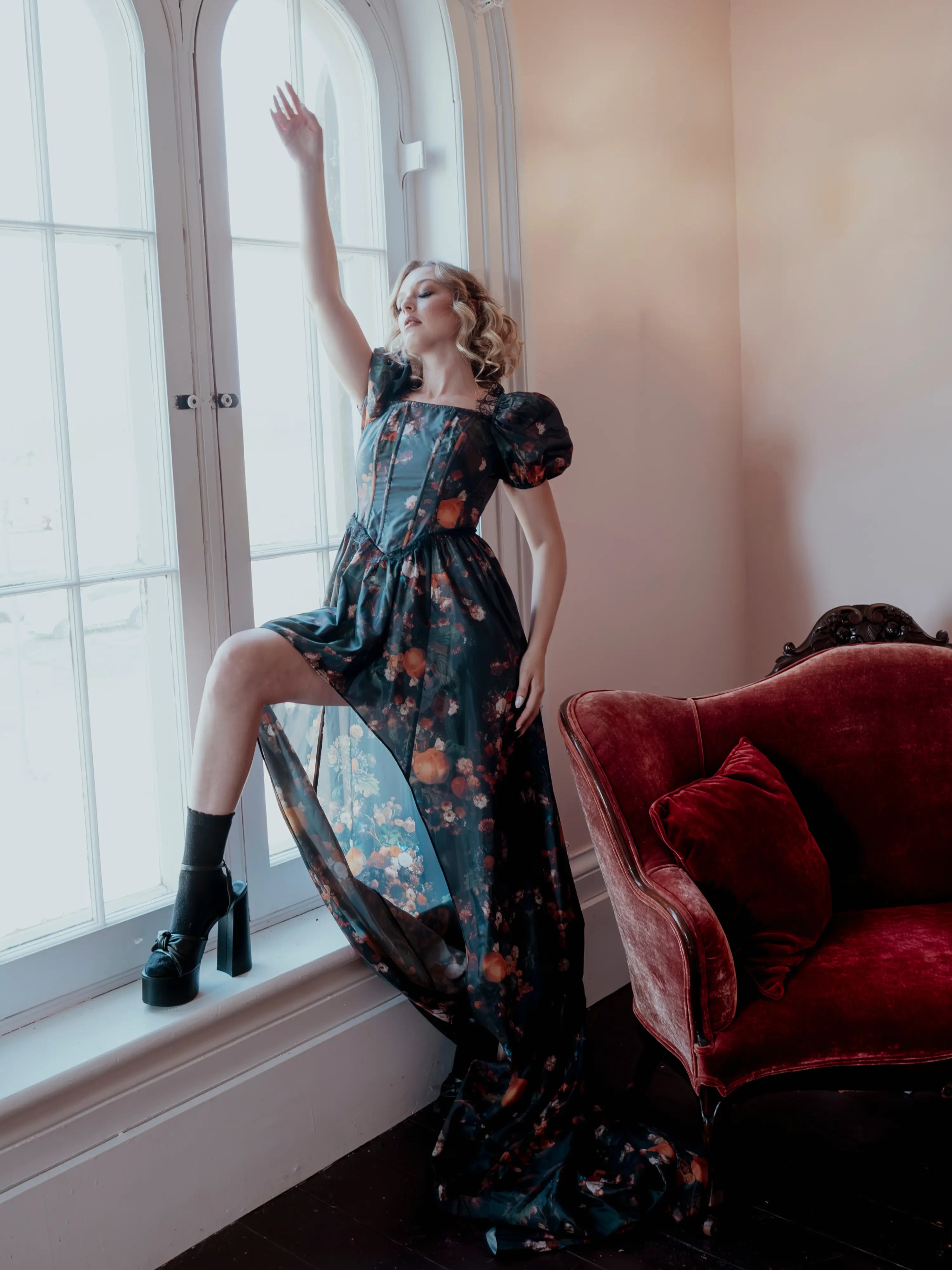 A woman wearing the still life printed taffeta dress is standing by a window with a red couch in the foreground.