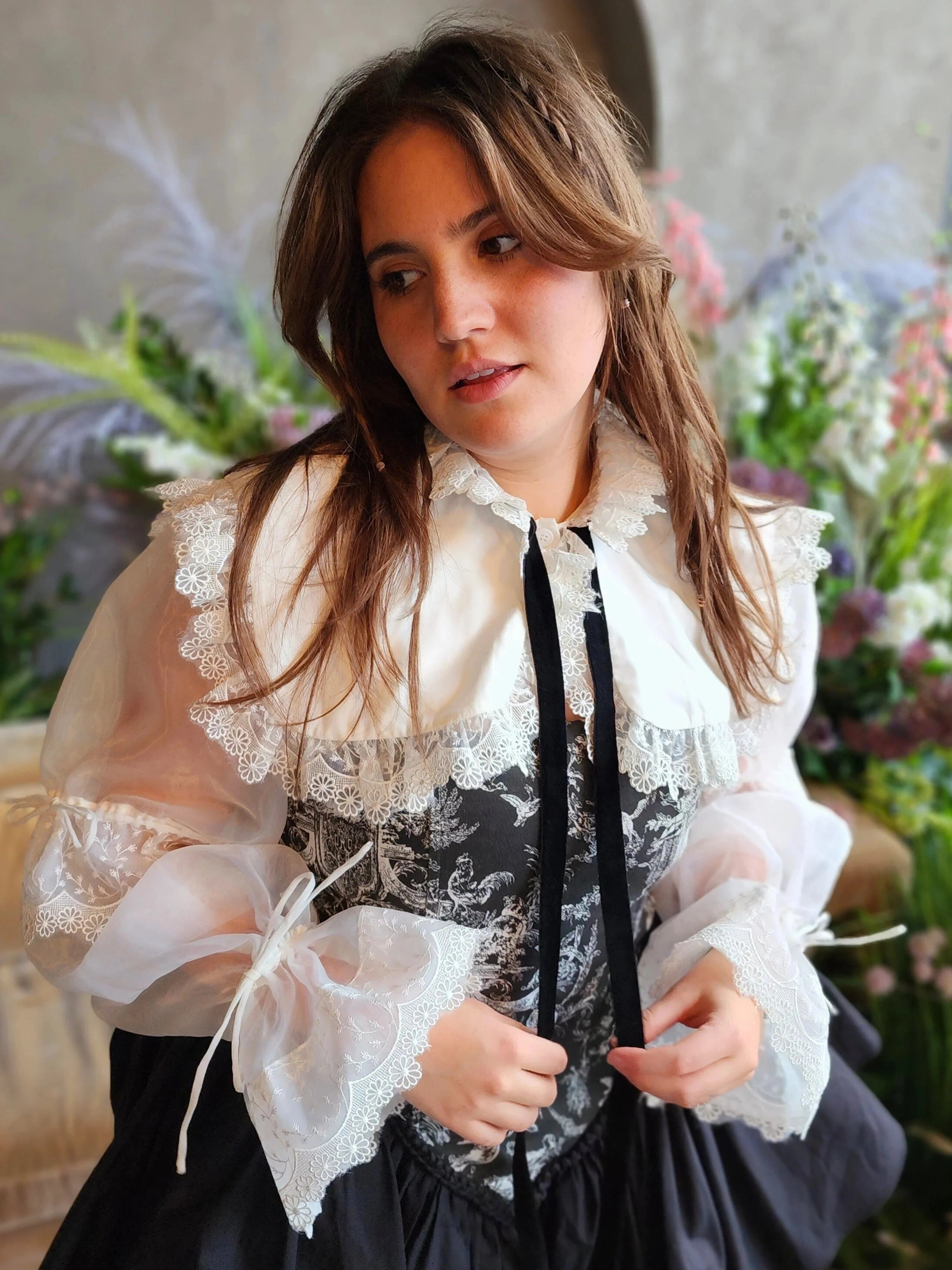 A woman in a ciela snow white shrug top featuring ornate lace trim and a black ribbon tie overtop a black corset mini dress.