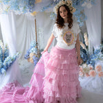 Front view of a woman wearing a t-shirt and a pink ruffle skirt in front of blue floral decorations.