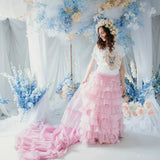 A woman in a white top with a kitten design and a pink ruffled skirt is standing in front of floral decorations.