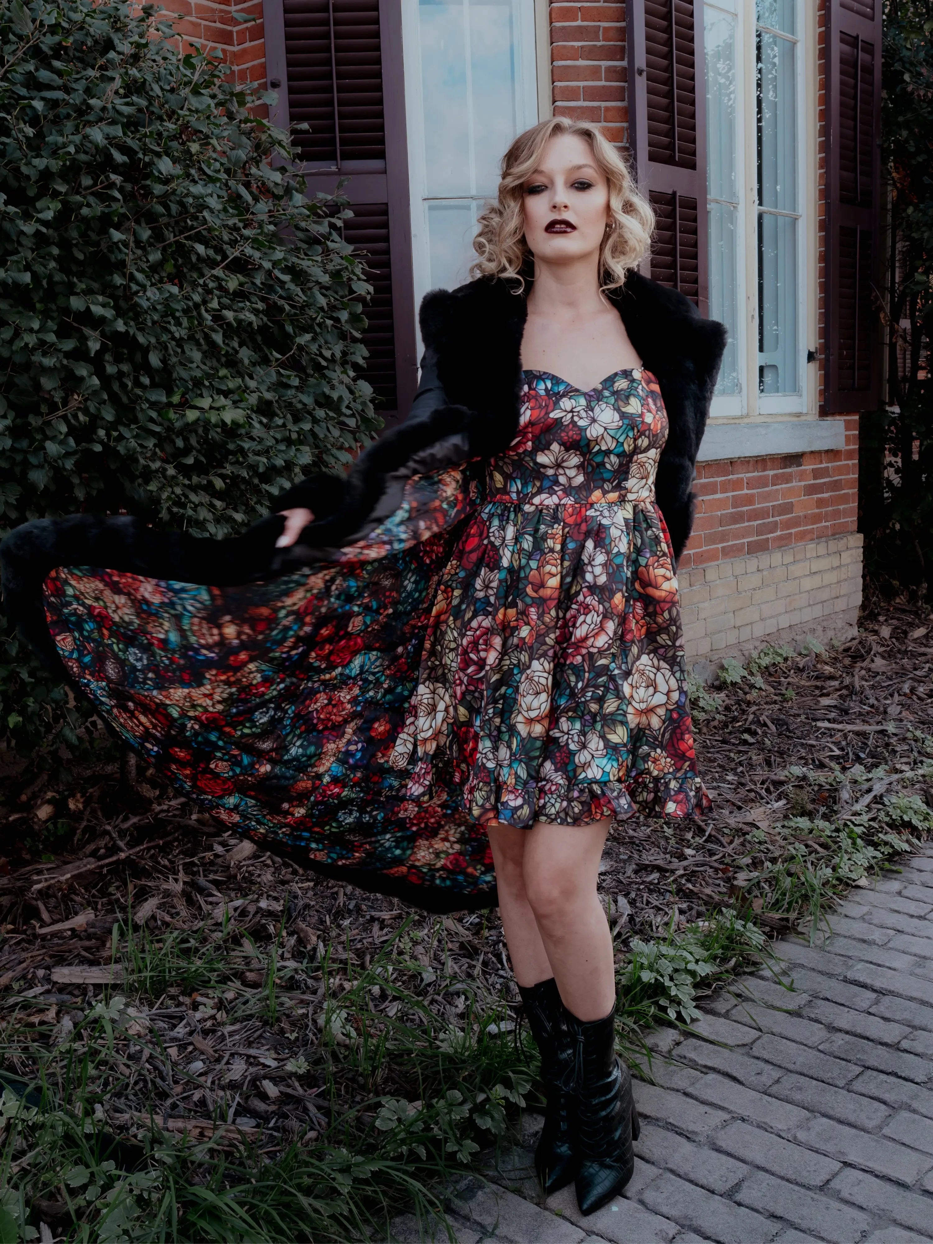 A woman wearing a stained glass satin mini dress and a black coat, standing outdoors near a building.