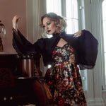 A woman wearing a stained glass satin mini dress and a black jacket, standing next to a piano in a room with large windows.