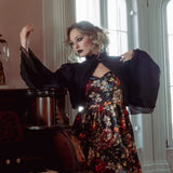 A woman wearing a stained glass satin mini dress and a black jacket, standing next to a piano in a room with large windows.