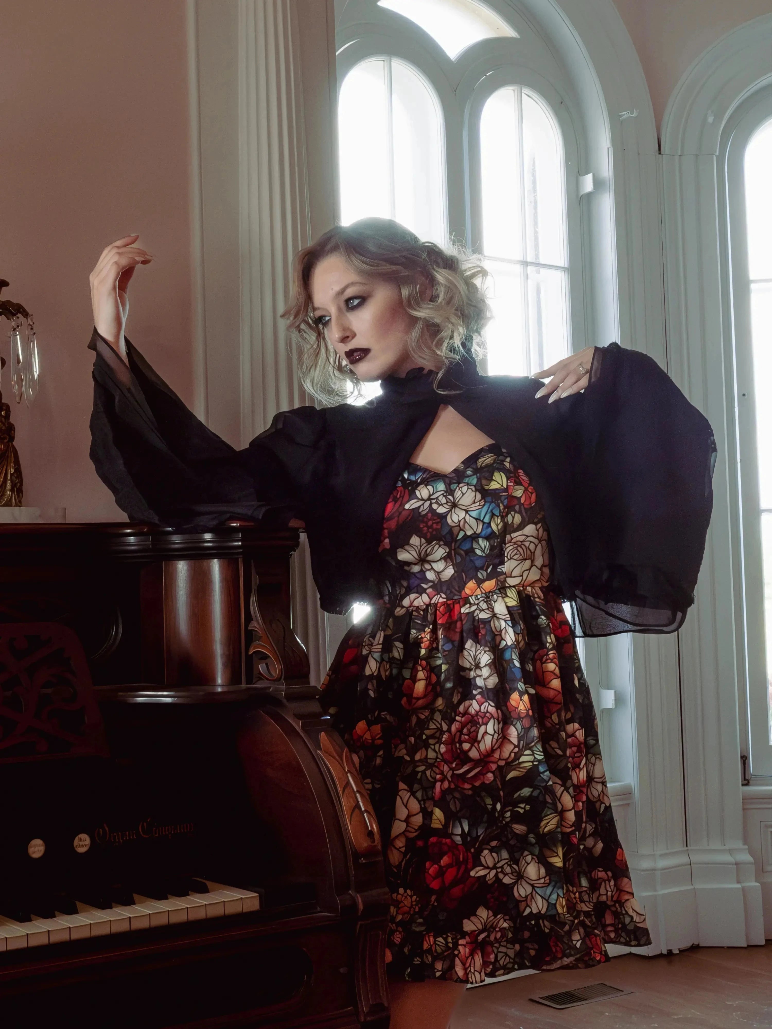 A woman wearing a stained glass satin mini dress and a black jacket, standing next to a piano in a room with large windows.