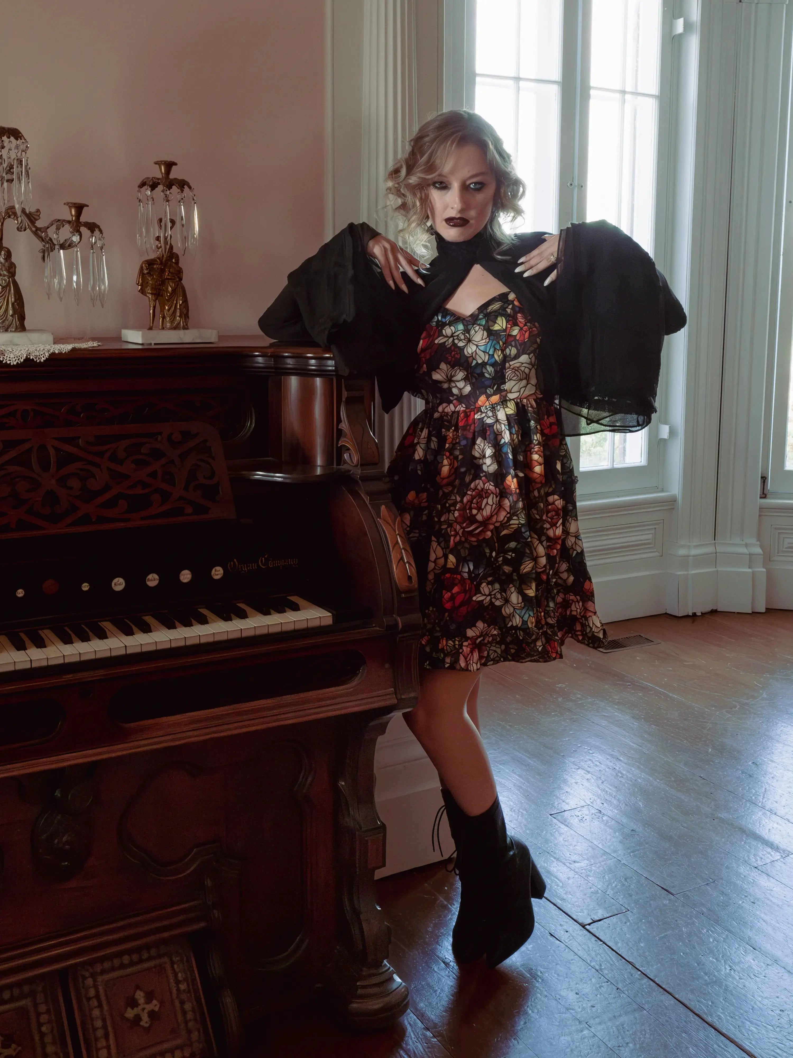 A woman wearing the Stained Rose Darling Dress, standing next to an old piano in a room with large windows.