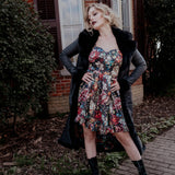 A woman wearing the Stained Rose Darling Dress and a black coat standing outdoors with a brick building and greenery in the background