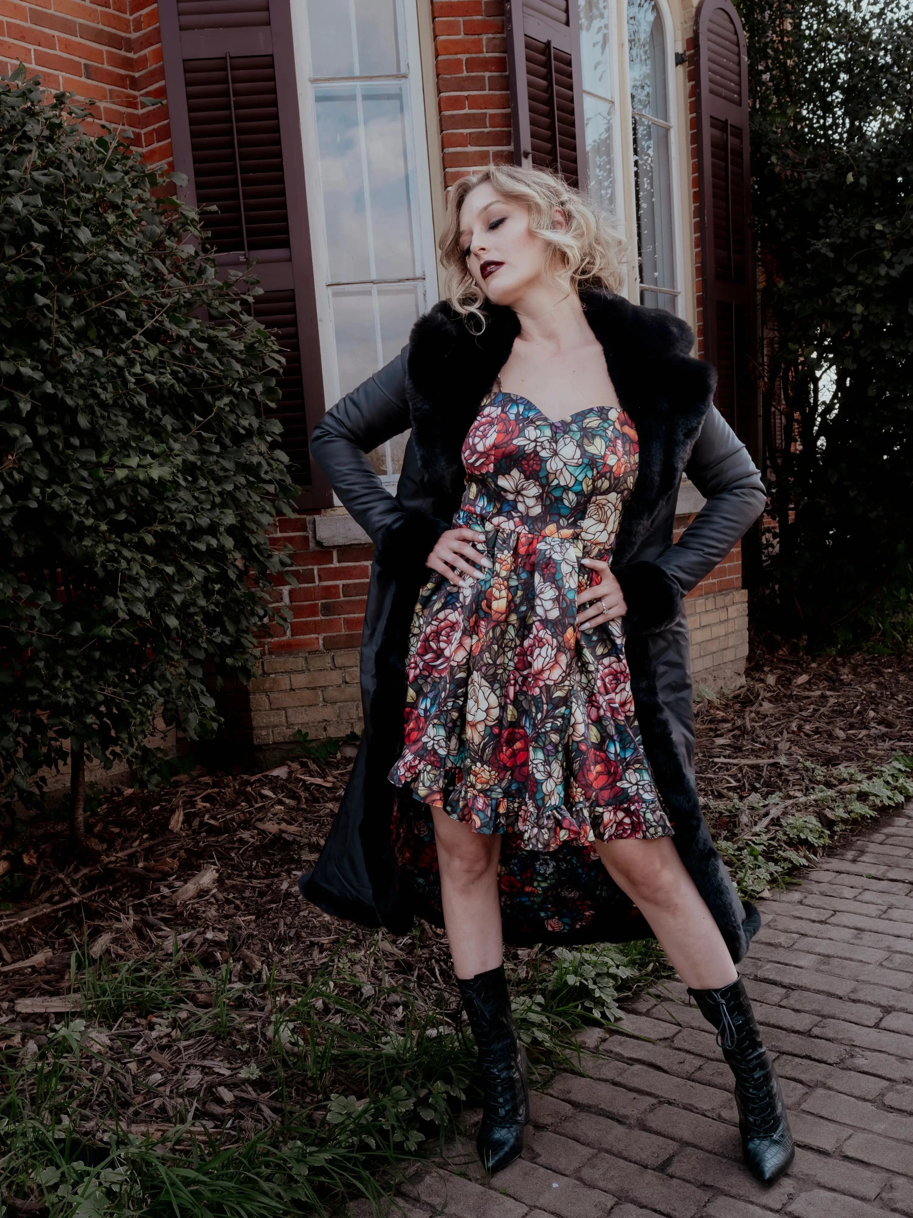 A woman wearing the Stained Rose Darling Dress and a black coat standing outdoors with a brick building and greenery in the background