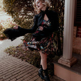 A woman wearing the Stained Rose Darling Dress and a black jacket, standing on a porch.