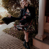 A woman wearing the Stained Rose Darling Dress and a black jacket, standing on a porch.