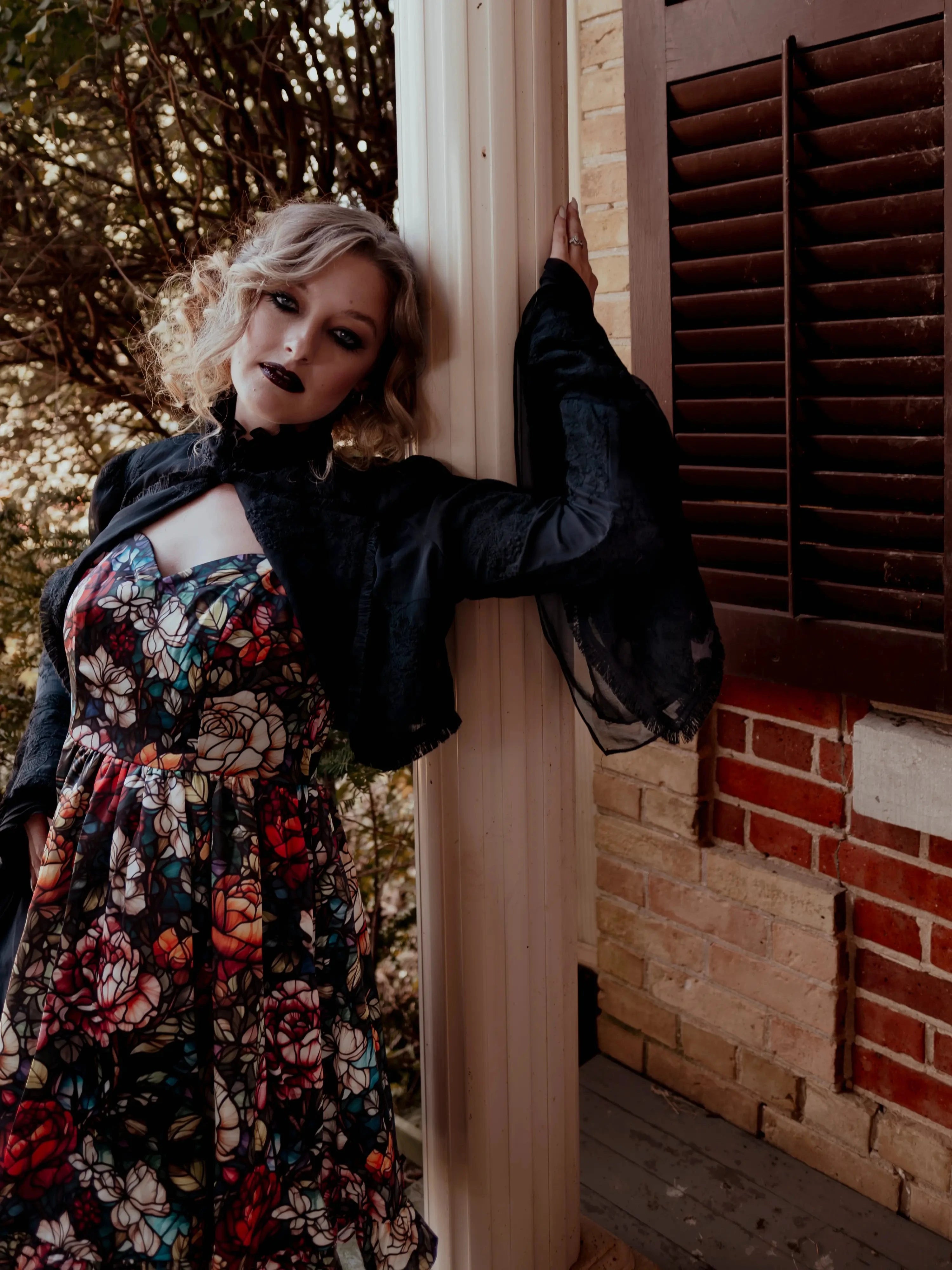 A woman wearing the Stained Rose Darling Dress, leaning against a brick wall with a window.