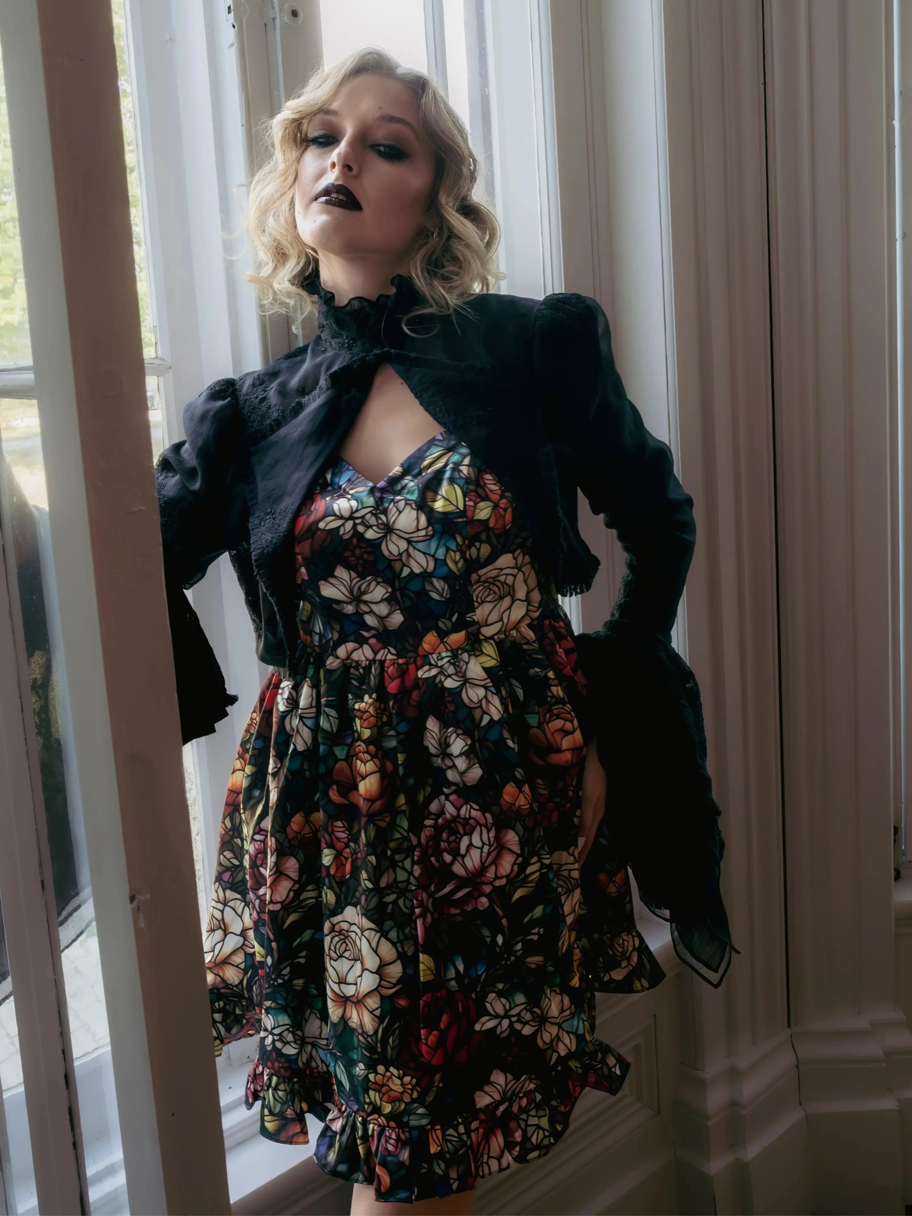 A woman wearing a stained glass satin mini dress and a dark jacket, standing in a doorway.