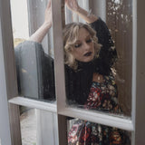 A woman wearing the Stained Rose Darling Dress, standing behind a window with raindrops.