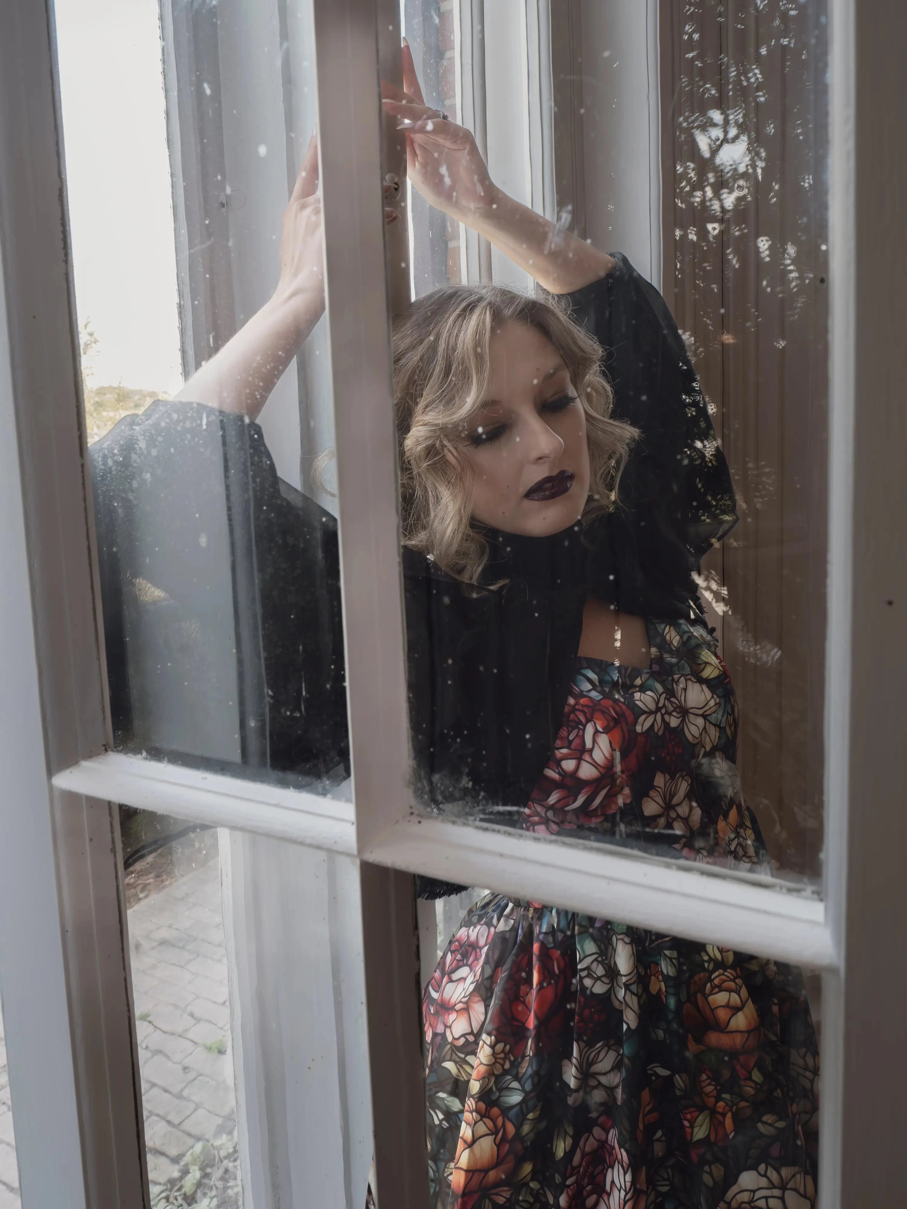 A woman wearing the Stained Rose Darling Dress, standing behind a window with raindrops.