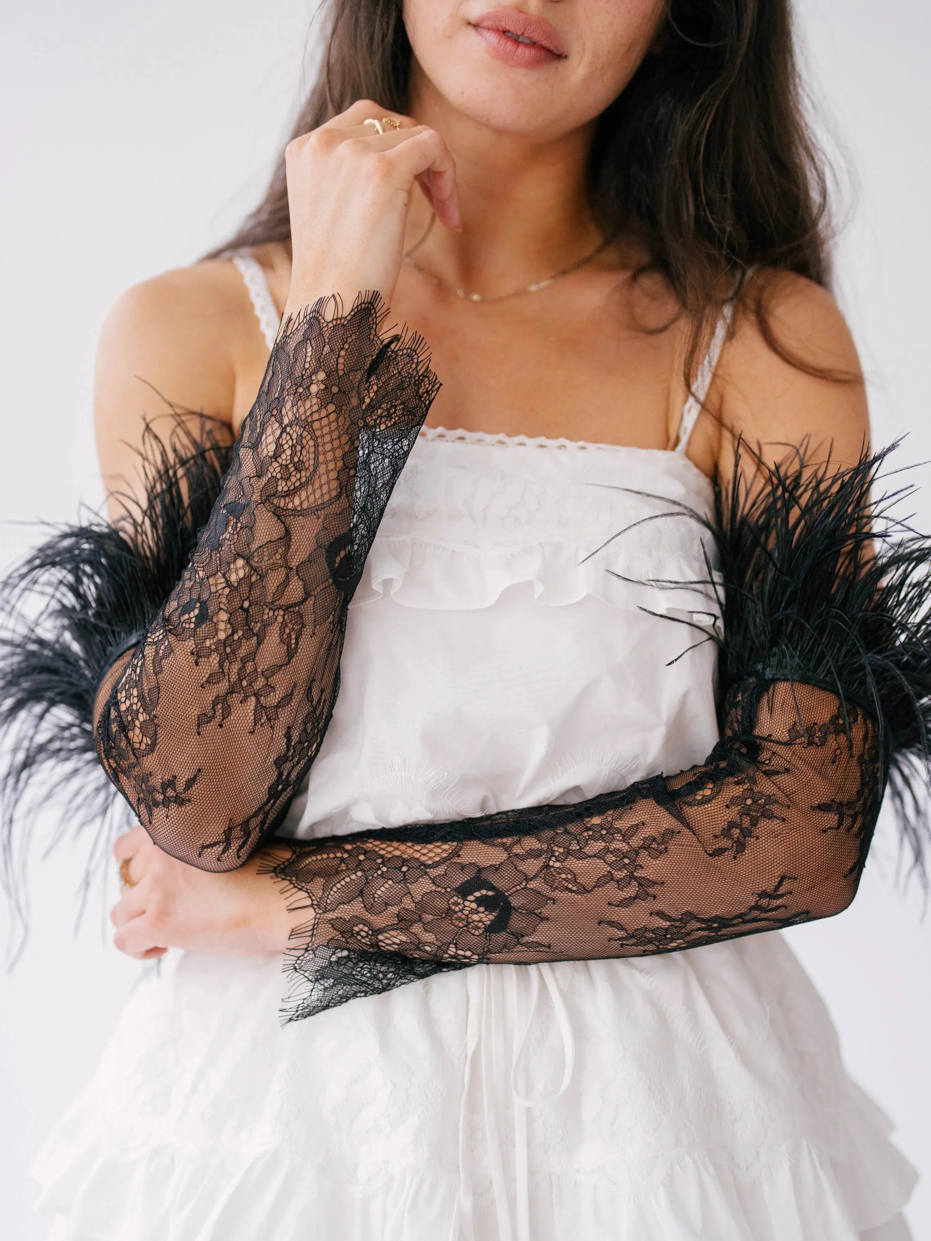 The Eleanor Black Feather Gloves featuring sheer black lace and dramatic feather trim, styled with a white ruffled dress for a bold contrast.