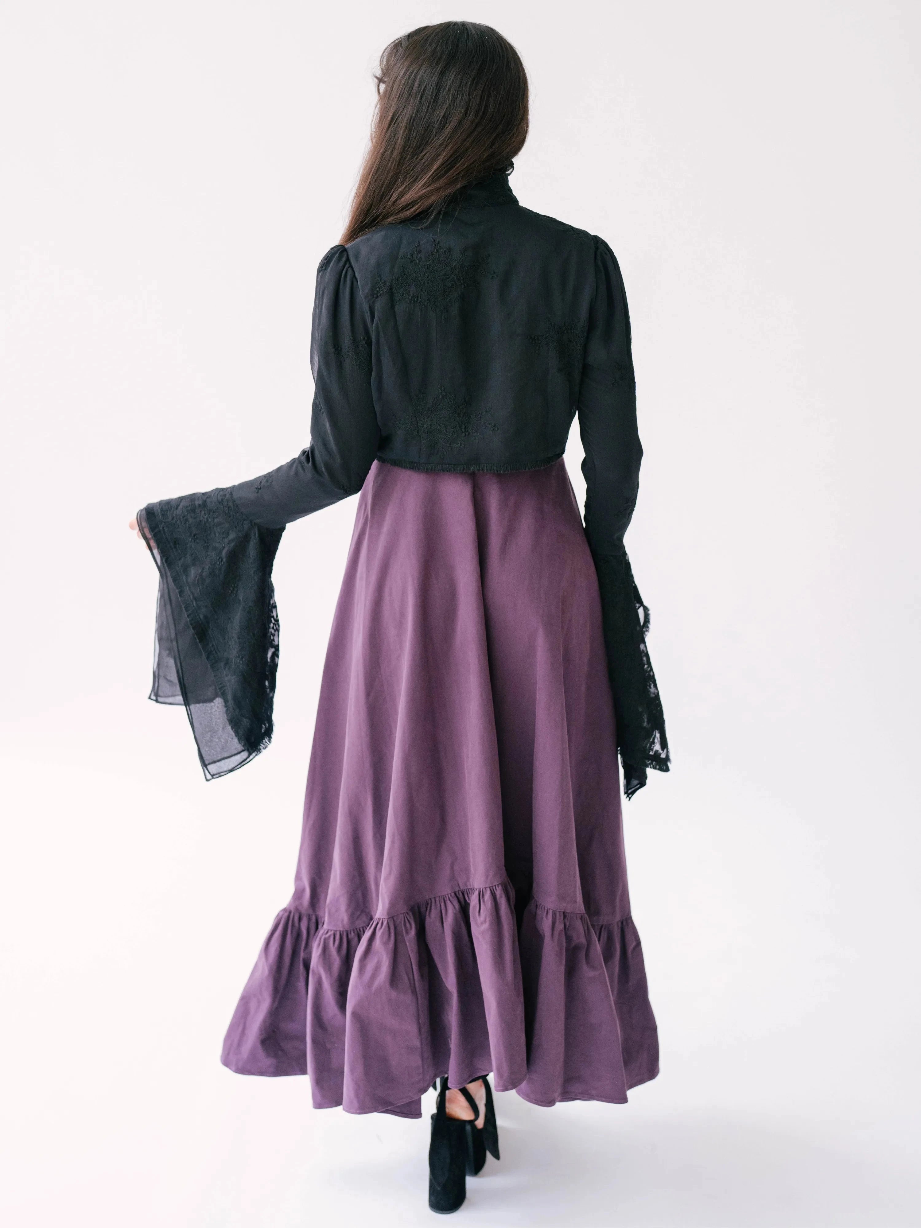 A back view of a woman wearing a purple midi dress with a black lace bolero jacket over top.