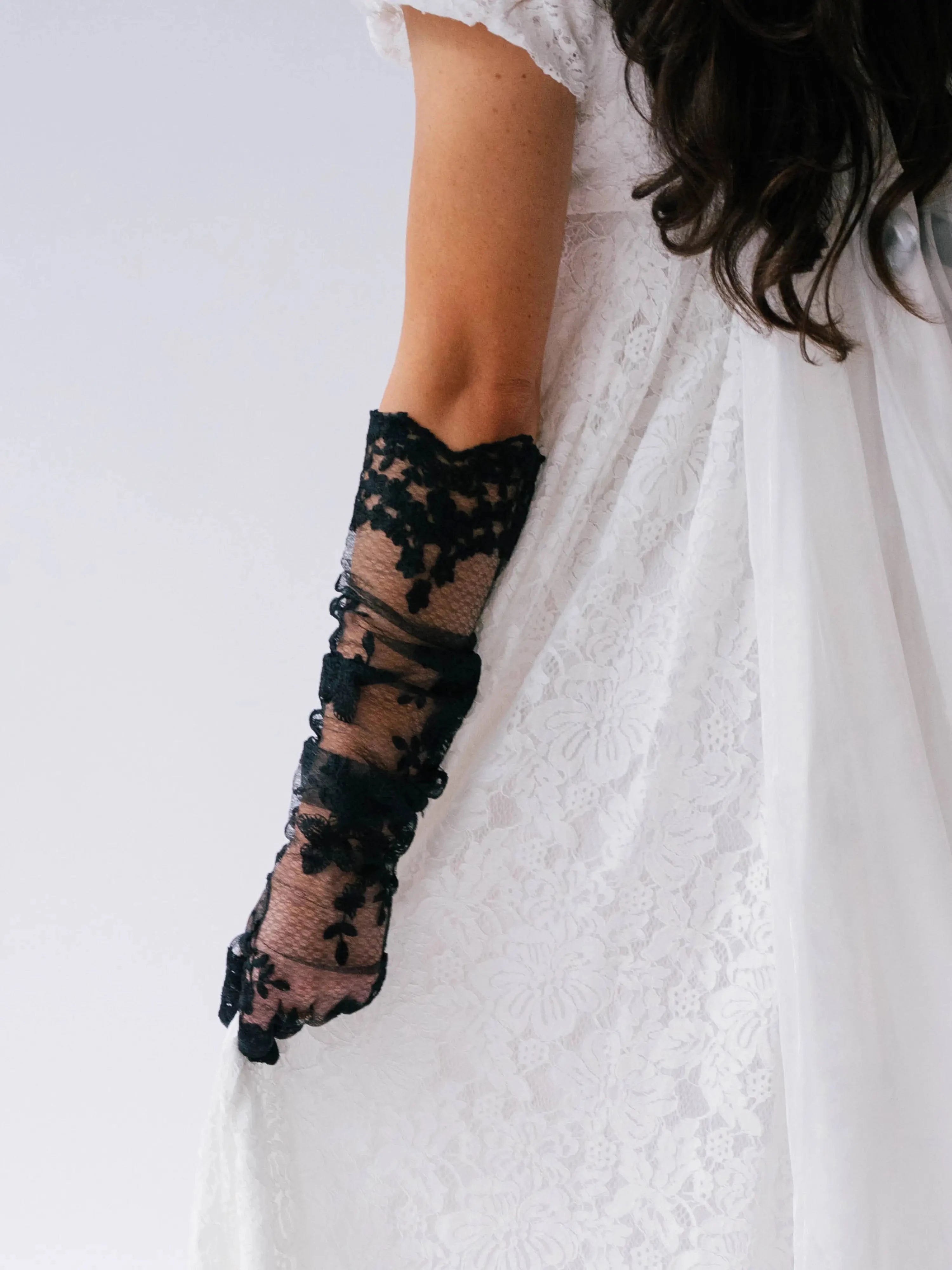 Black lace opera gloves worn with a white dress on a plain background