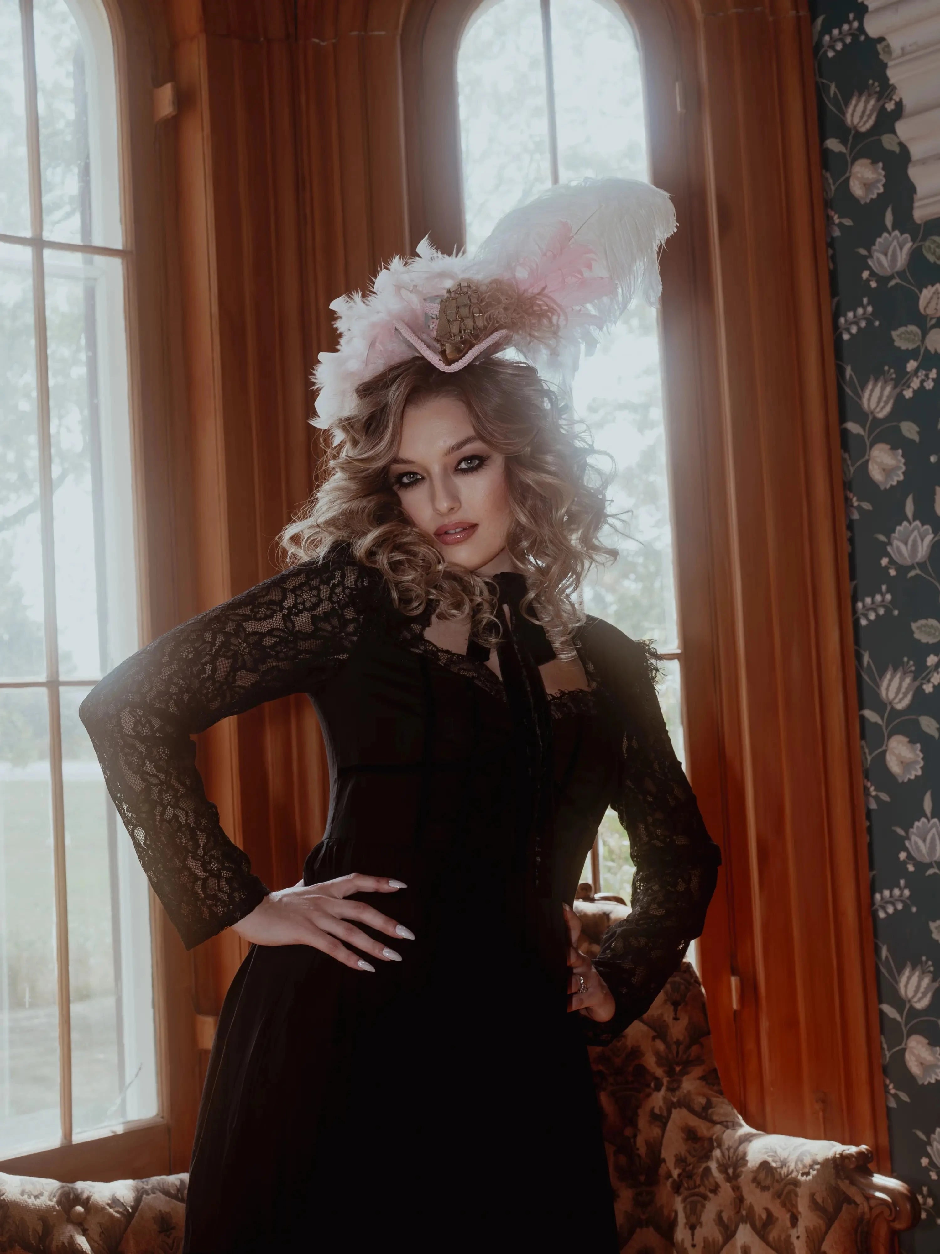 A woman in a black cotton dress with a decorative hat in a room with floral wallpaper.