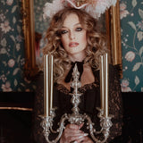 A woman in a black cotton dress holding a silver candelabra in front of a decorative mirror with floral wallpaper.