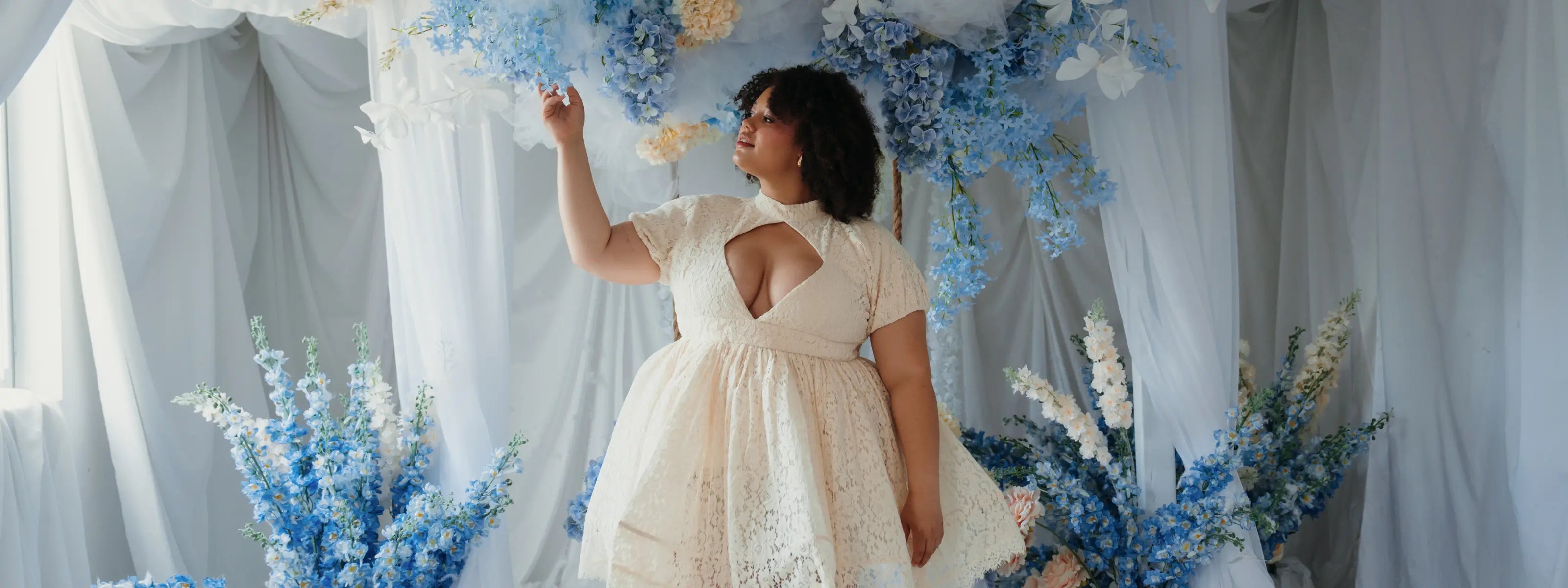 Woman in a white dress standing among blue floral decorations with sheer curtains in the background